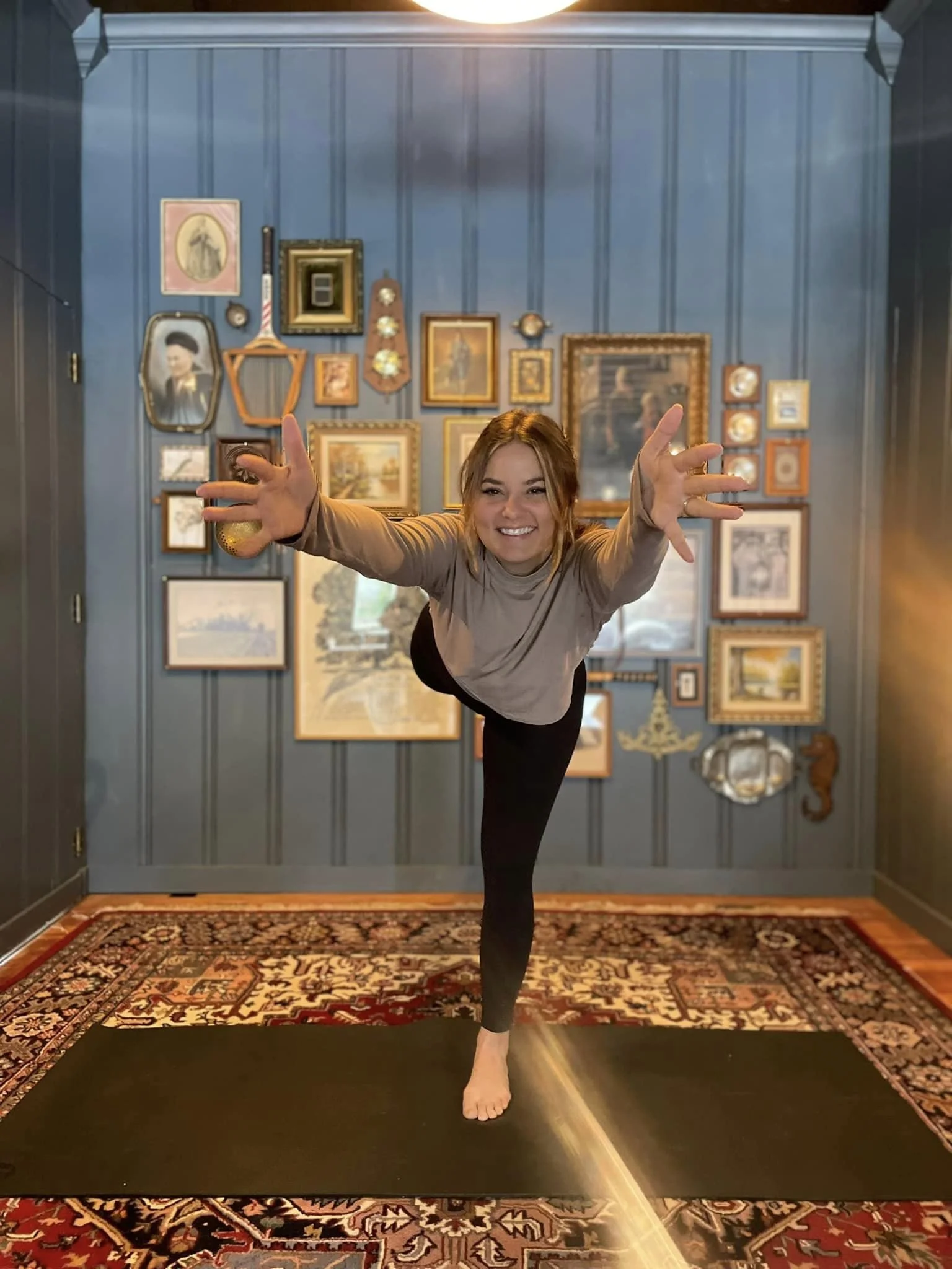 A woman practicing yoga or dance on a black mat in a room with blue walls decorated with various picture frames and artwork.