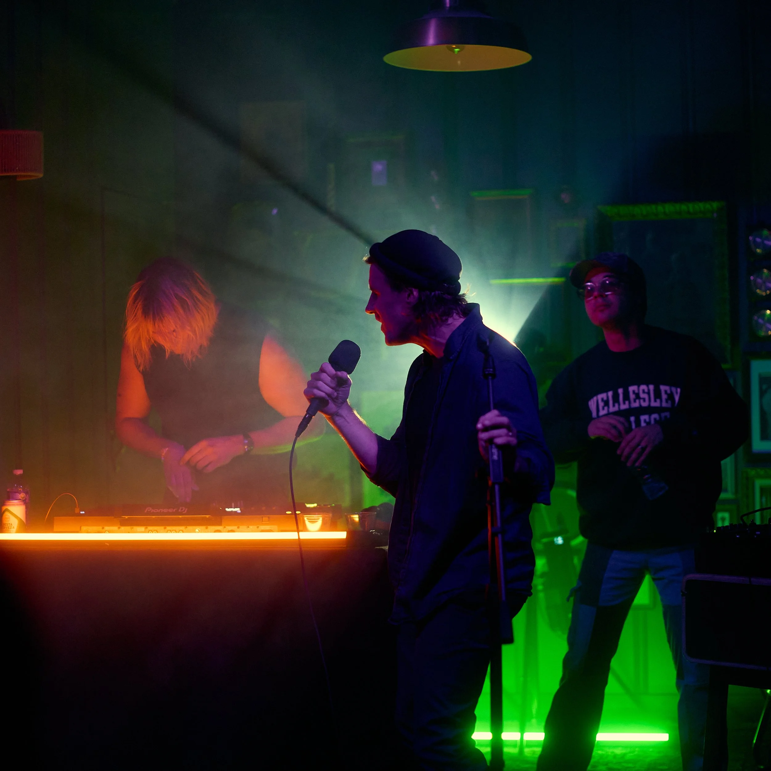 People performing on stage in a club with colorful lighting and a DJ setup.