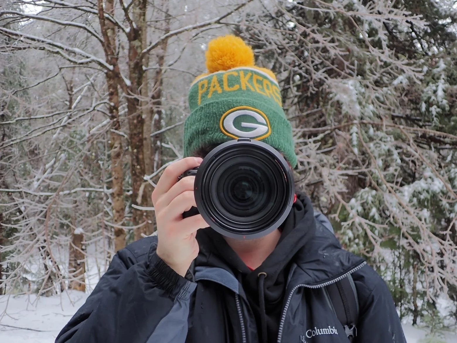 Person in winter clothing taking a photo with a camera in a snowy, wooded area. The person wears a Green Bay Packers beanie with a yellow pom-pom, and a black jacket.