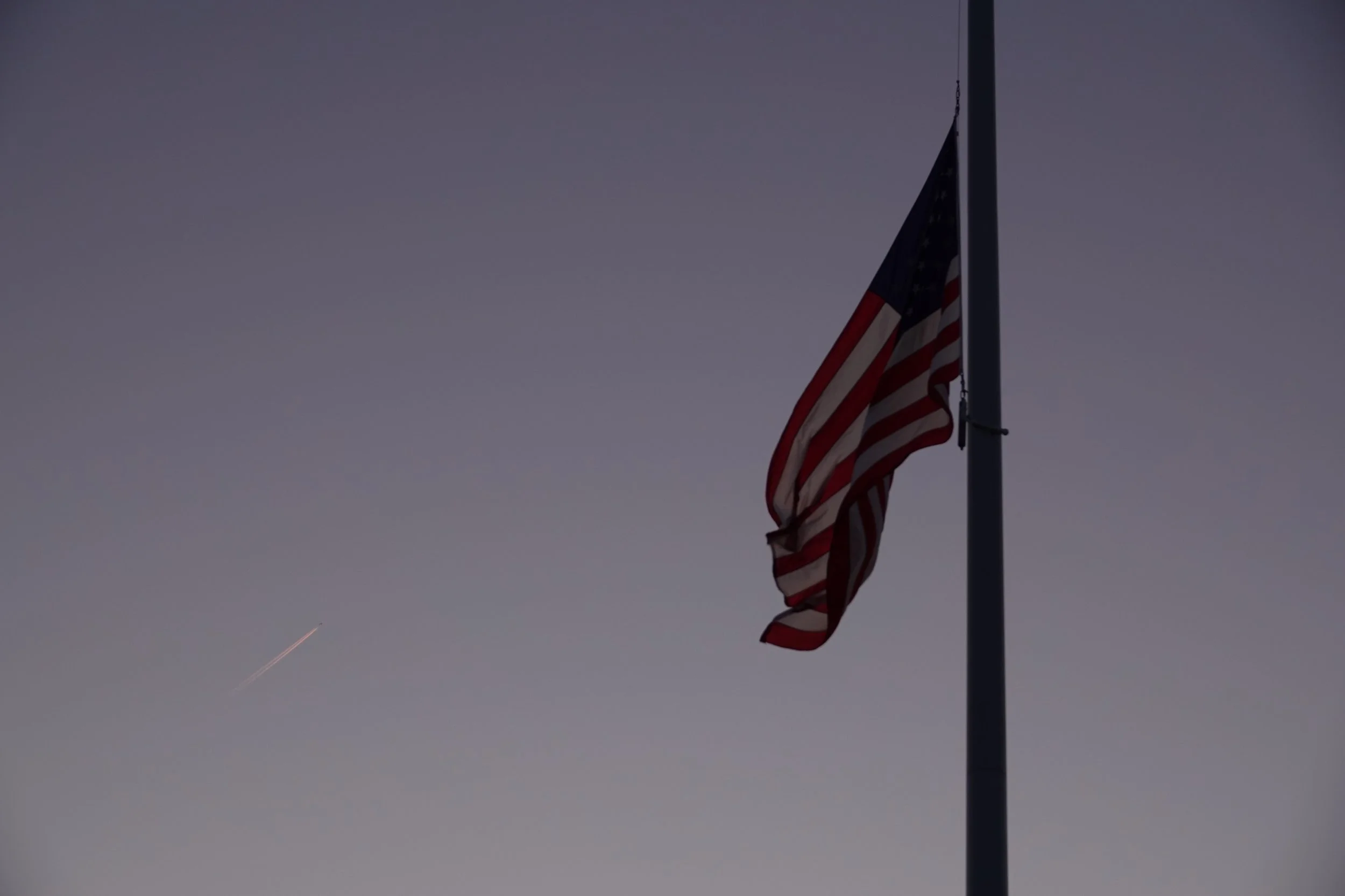 An American flag on a flagpole against a clear sky with a faint jet trail in the background.