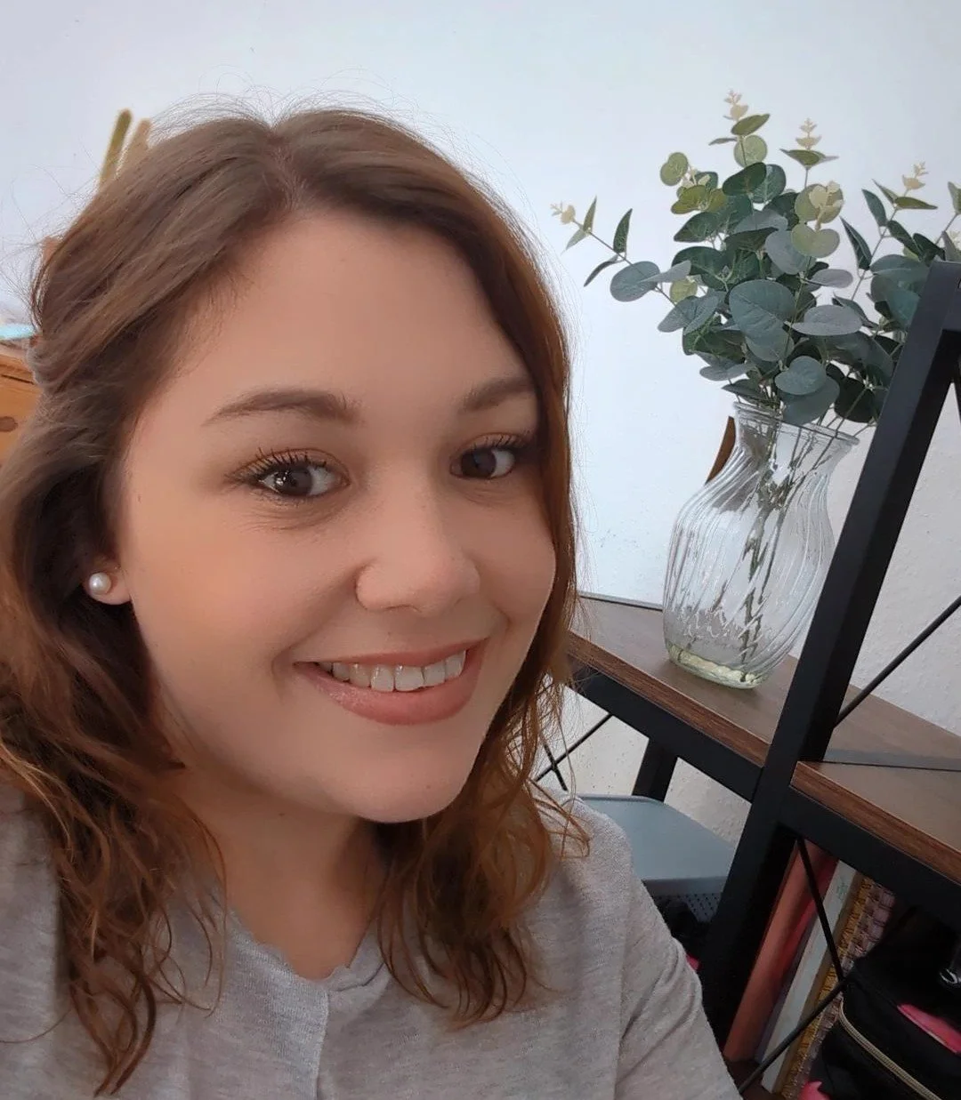 A young woman with curly brown hair, smiling at the camera, sitting indoors with a wooden desk, a clear glass vase with green eucalyptus leaves, and a black metal shelf in the background.