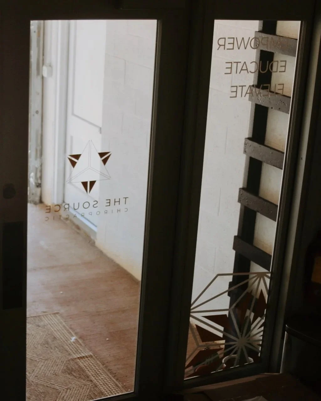 Interior view of a glass door with the logo and name 'The Source Chiropractic'. Part of a staircase and a wall are visible outside through the glass.