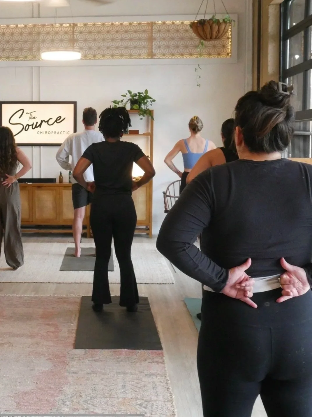 People participating in a yoga or fitness class in a studio with wooden furniture and a sign that reads 'The Source Chiropractic' on the wall.