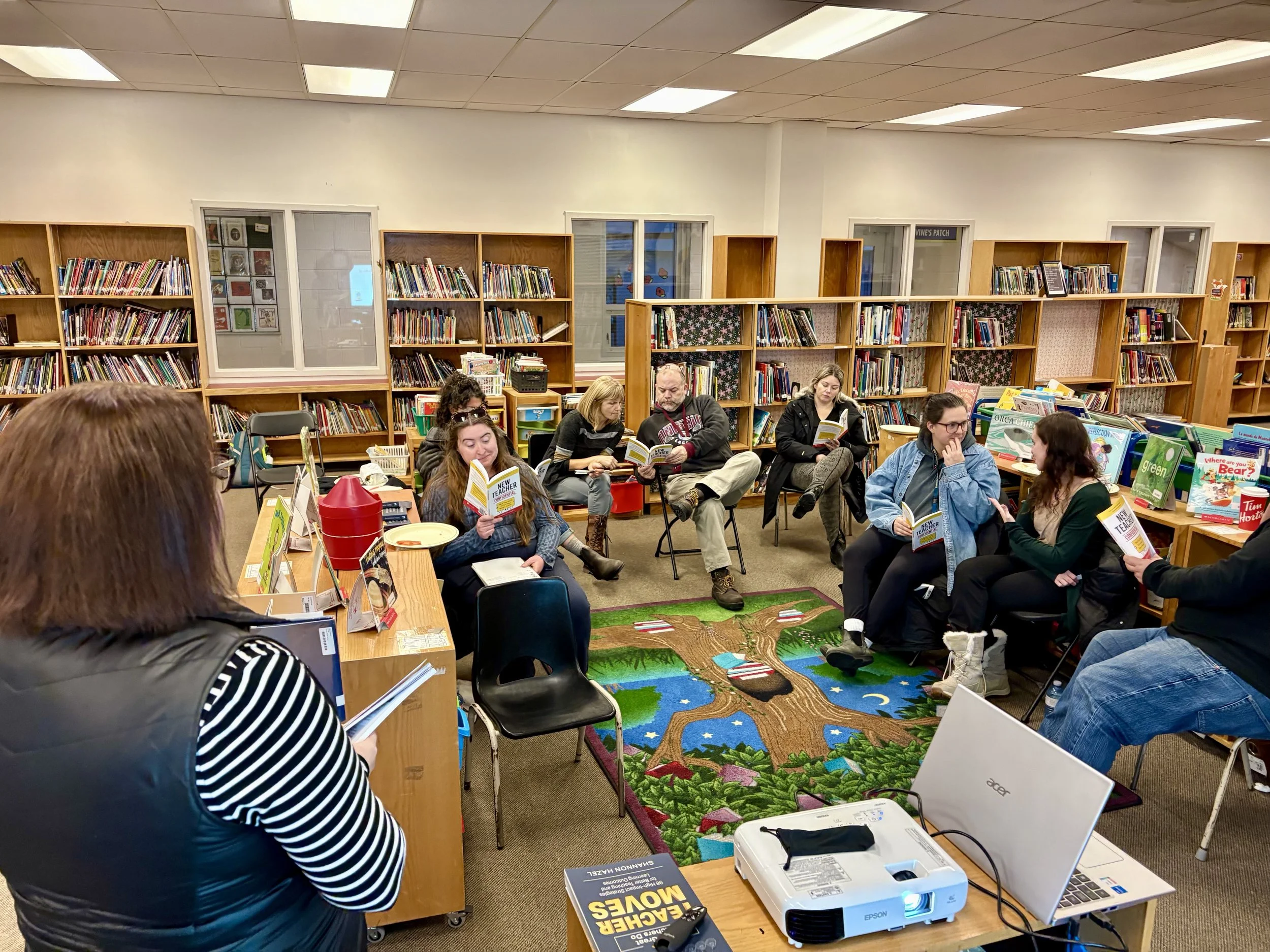 People sitting in chairs in a library reading books. Bookshelves filled with books are visible behind them, and there is a colorful carpet on the floor depicting a landscape with trees, rivers, and animals. People's faces are visible as they focus on