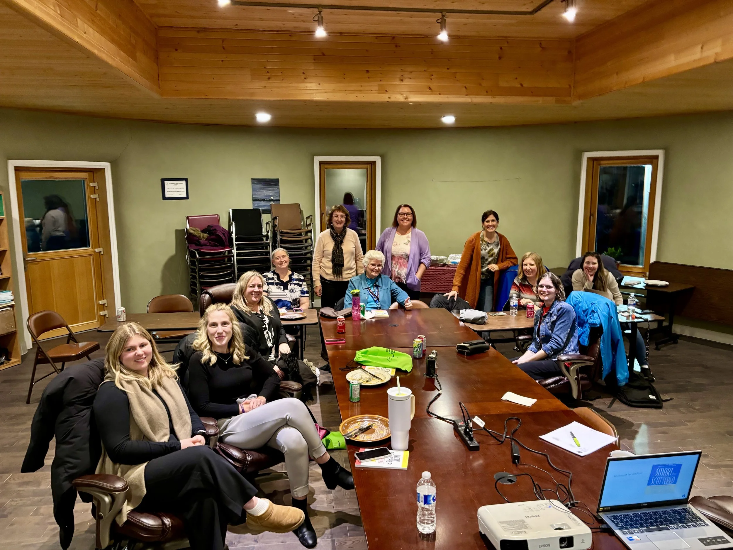 Group of women gathered around a large conference table in a room with green walls and wooden ceiling, smiling at the camera. The table has drinks, notebooks, and electronics, with chairs stacked against the wall and a window showing a nighttime scen