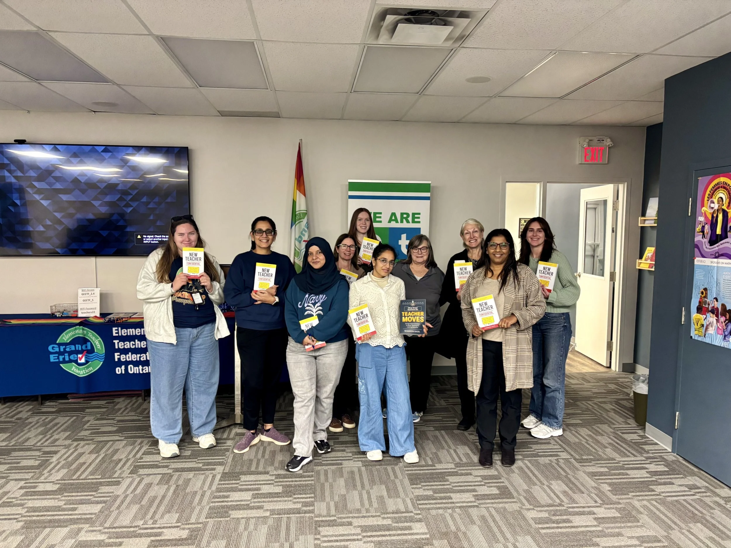 Group of fifteen people, mostly women, standing in a room holding books titled 'New Teacher'. They are smiling and posing for the photo. The background shows a large flat screen, a banner with the logo 'Grand Erie District School Board' and the words
