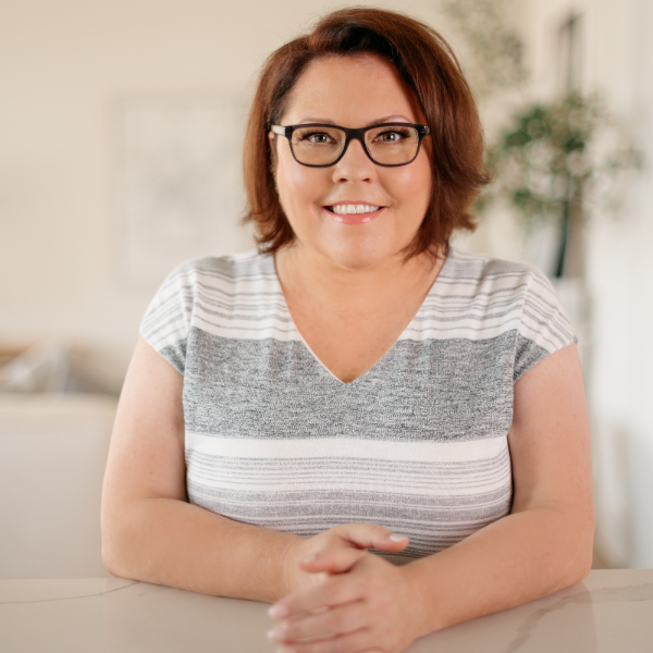 Shannon Hazel--A woman with short brown hair, wearing glasses and a striped gray and white top, sitting at a table and smiling in a well-lit room.