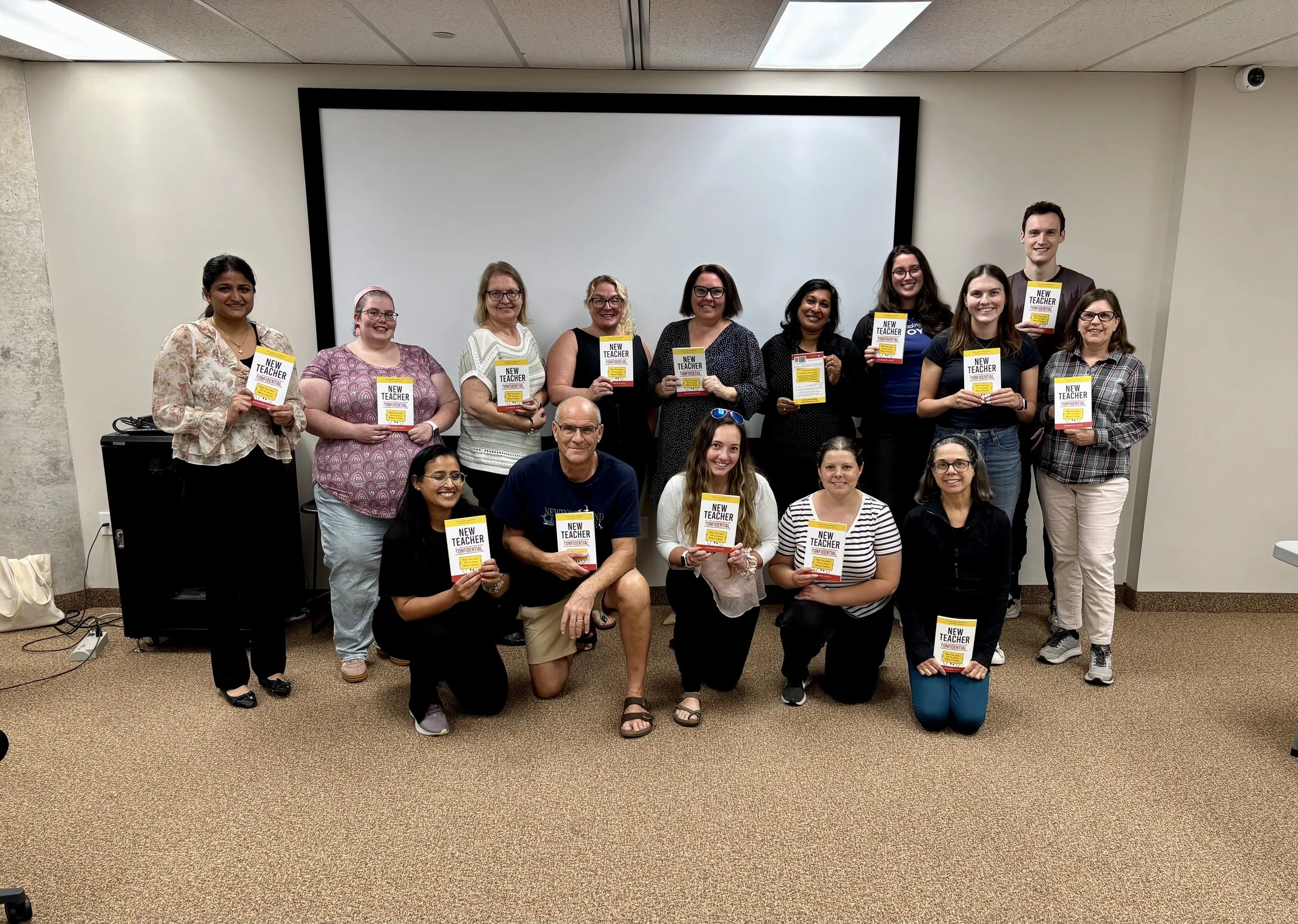Group of people, mostly women, standing and kneeling in front of a presentation screen, holding books titled "New Teacher Confidential." They are smiling in a classroom or conference room setting.