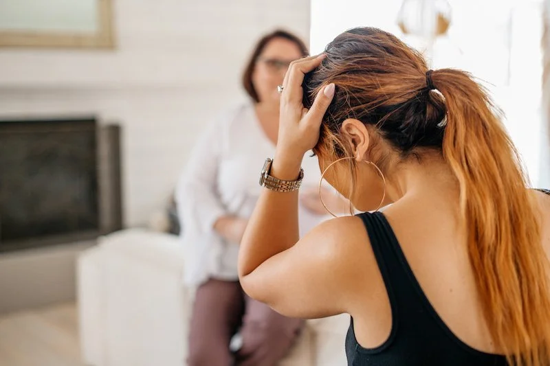 Shannon supporting a teacher. Young woman with red hair holding her head in distress, sitting in a room with a woman in the background.