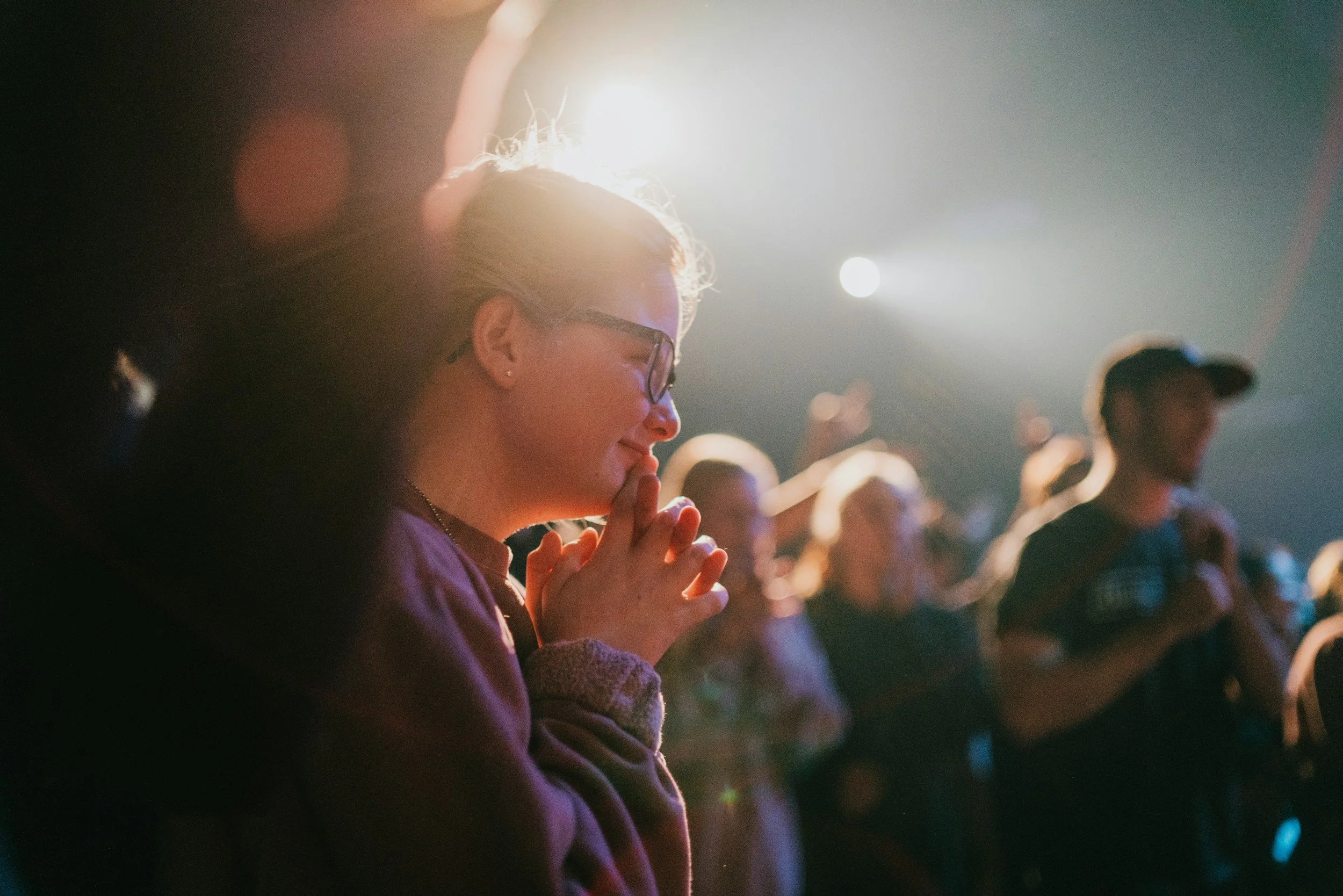 A woman with glasses and earrings praying with her hands clasped during a religious gathering, with others in the background, illuminated by stage lights.
