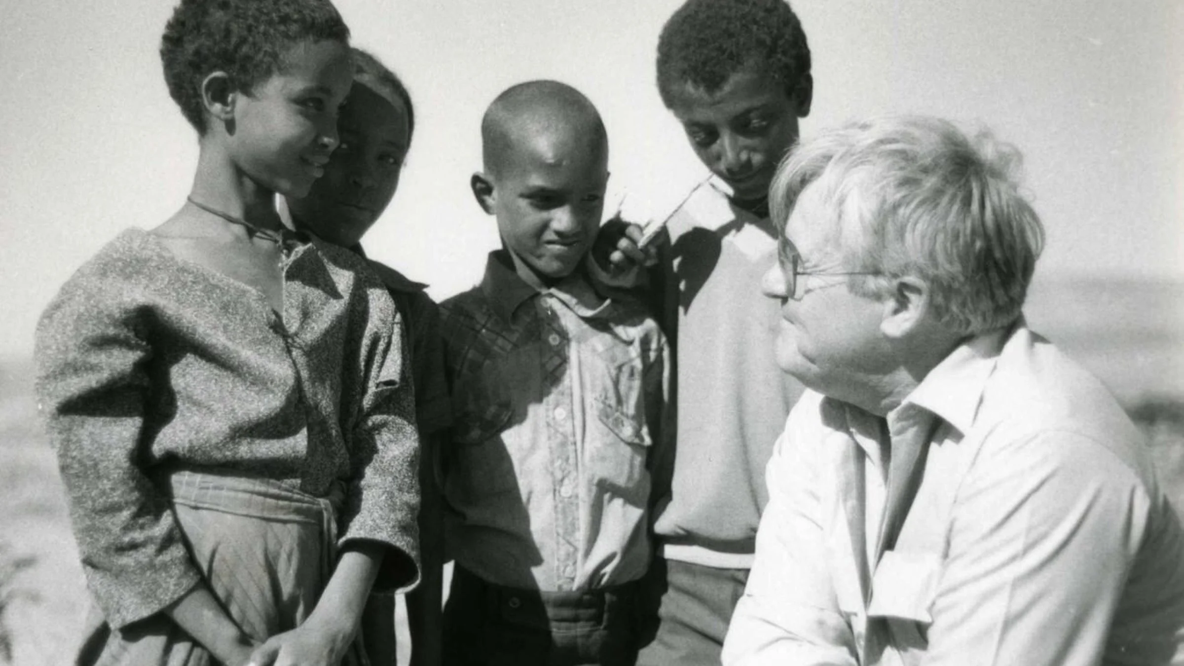 A group of five people, four children and an older woman, are standing outdoors engaging in conversation. The children appear to be listening intently to the woman.