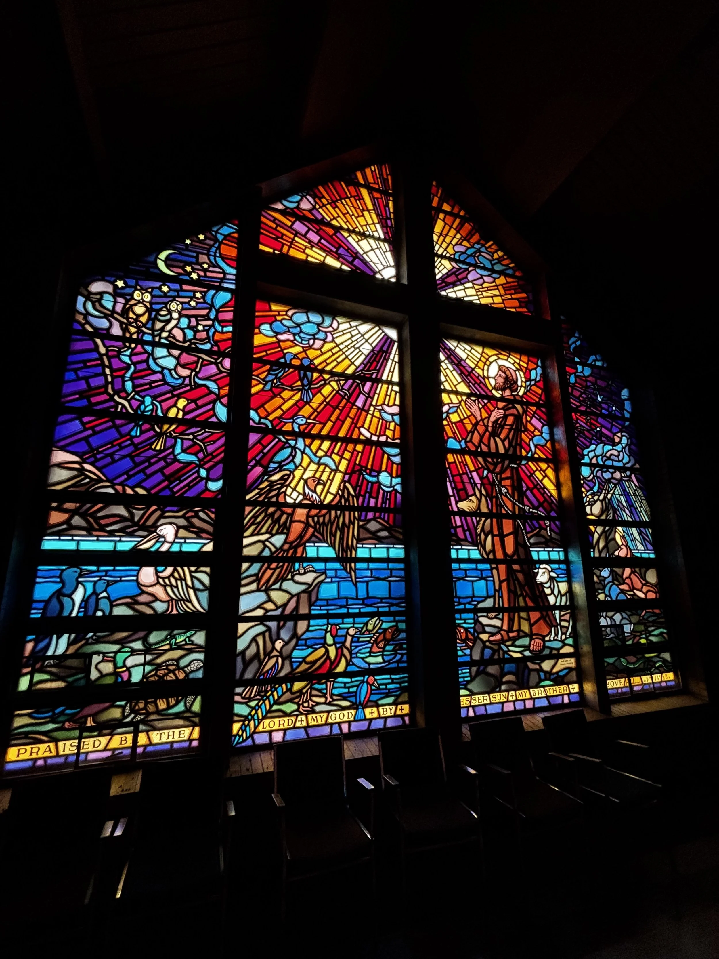 Colorful stained glass window depicting biblical scenes, with rays of light in the background and religious figures, birds, and text prominently displayed.