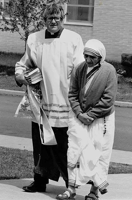 A woman dressed in religious attire, holding a book and some cloth, walking alongside an elderly woman wearing a headscarf, outside near a brick building.