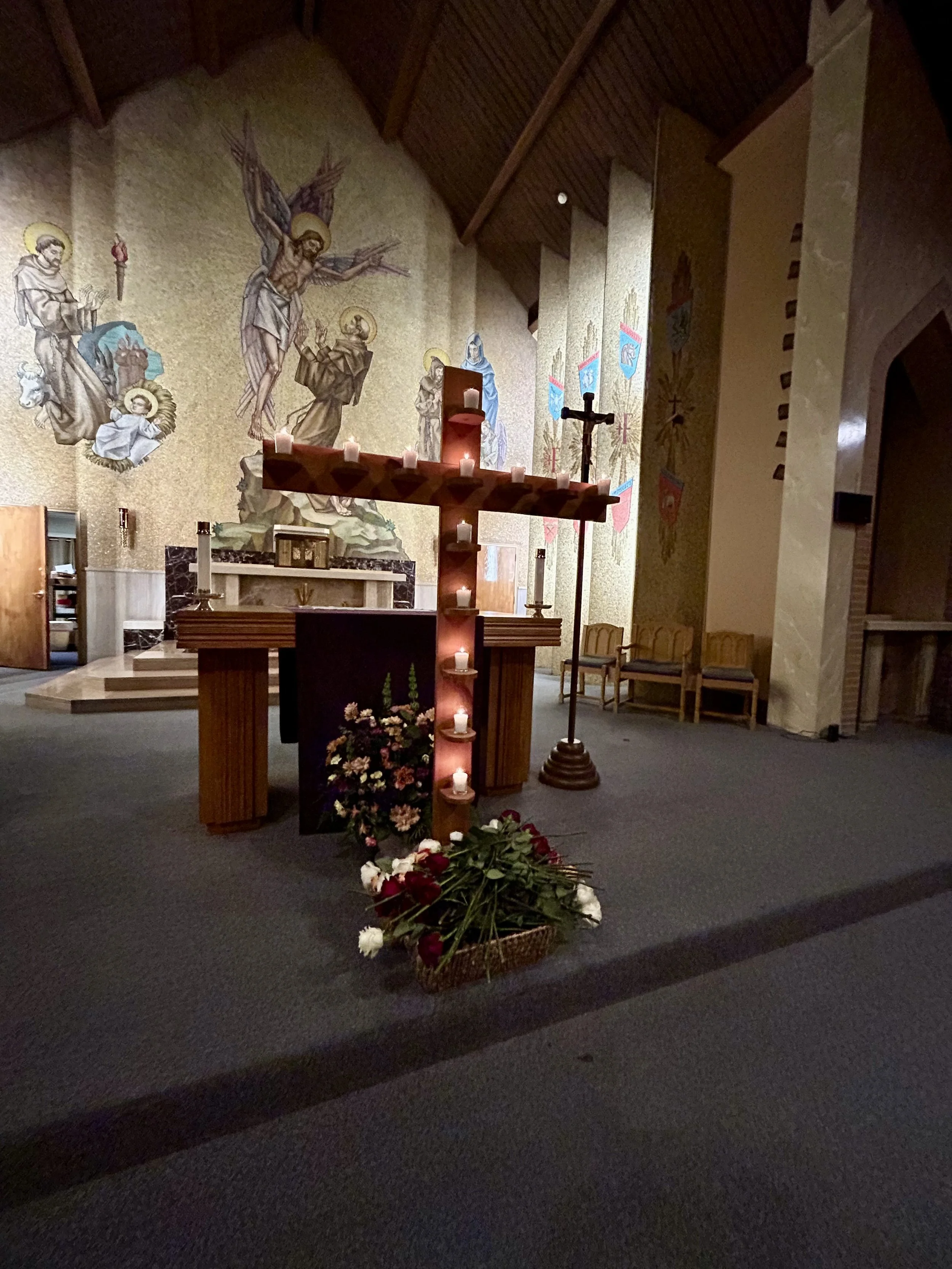 Chapel of St. Francis of Assisi.  The interior of a church altar with a large mosaic mural depicting Jesus Christ's crucifixion, surrounded by St. Francis of Assisi receiving the Stigmata. Candles and flowers are placed at the altar.