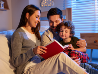 mother, father, and son sitting next to bed and reading a book