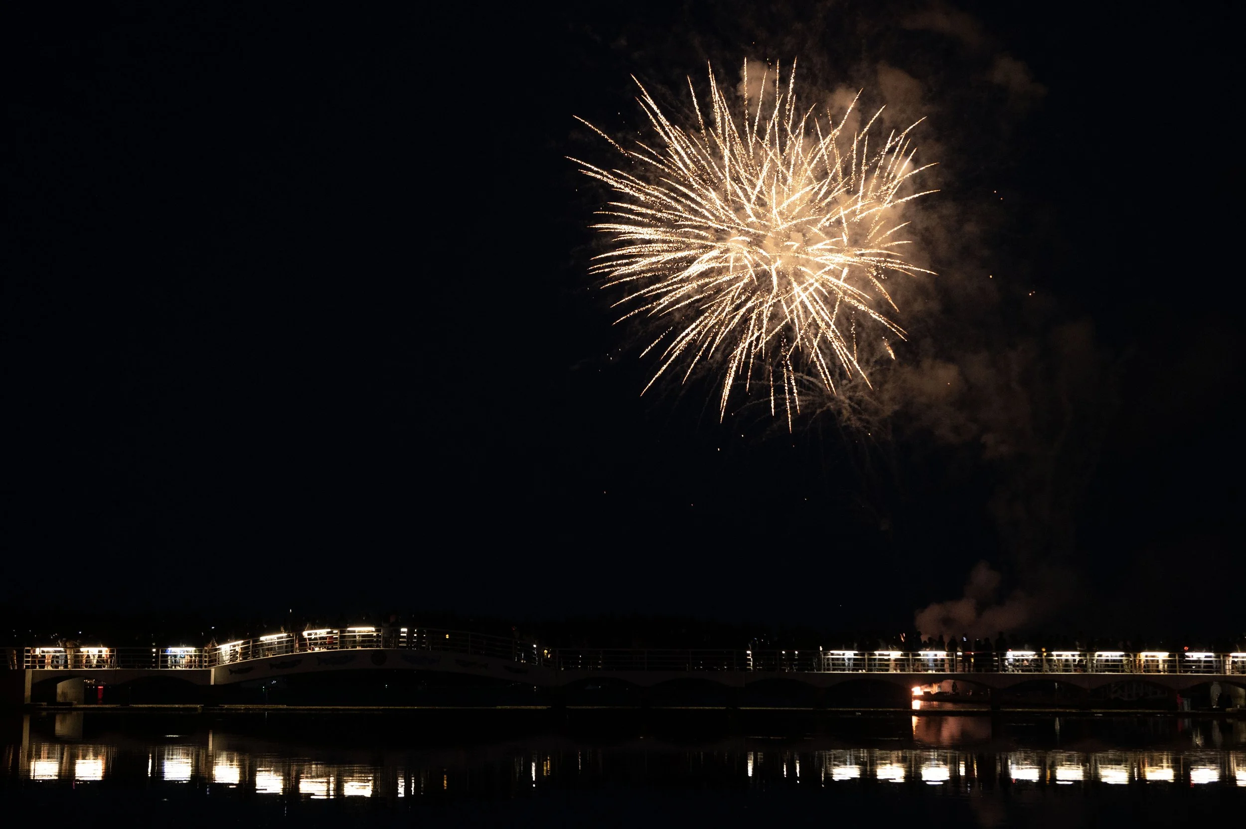 Fireworks display in the night sky above a bridge over water, with reflections visible below.