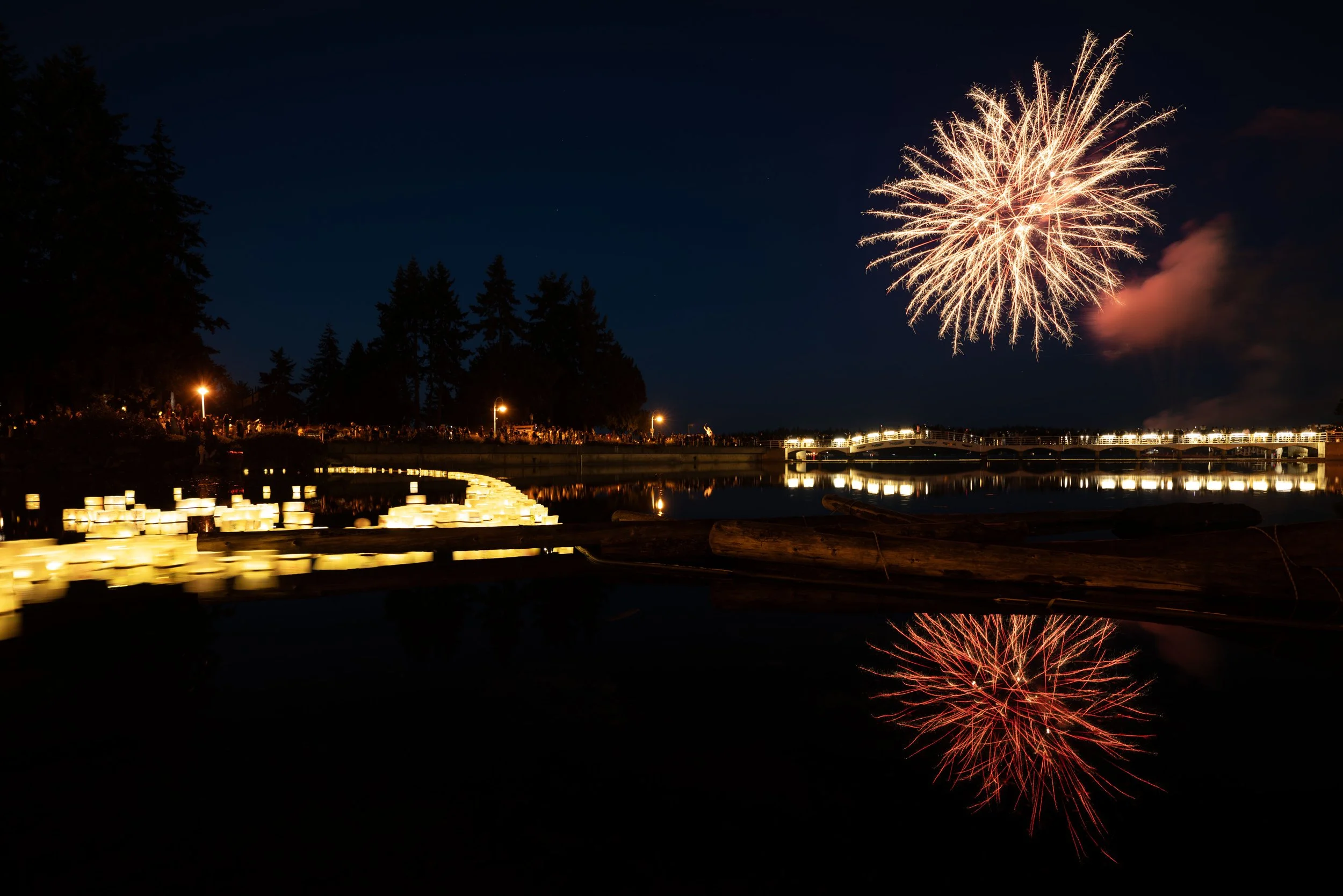 Fireworks explode in the night sky above a crowd of people gathered near a waterway, with reflections on the water and a string of illuminated candles or lanterns along the shoreline.