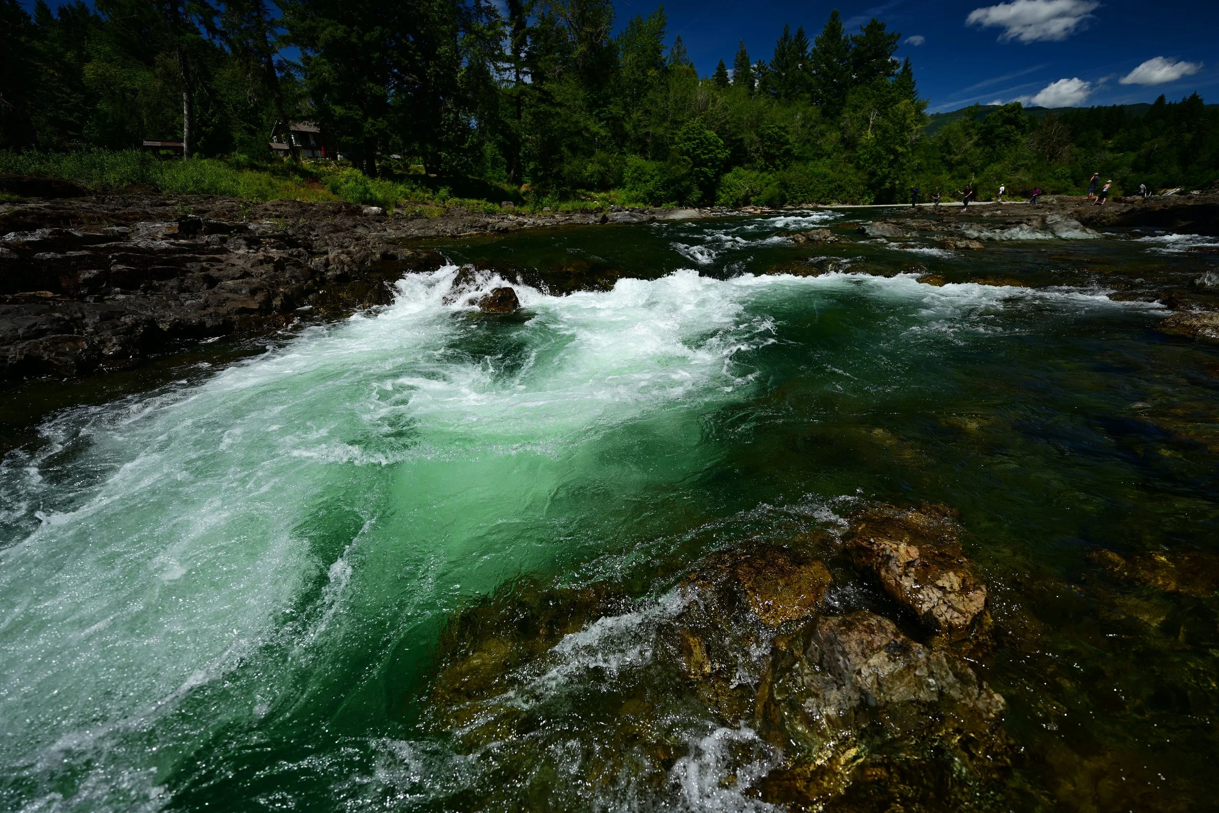 A river flowing with white water rapids, surrounded by rocks and greenery, with people fishing along the riverbank and forested hills in the background under a partly cloudy sky.