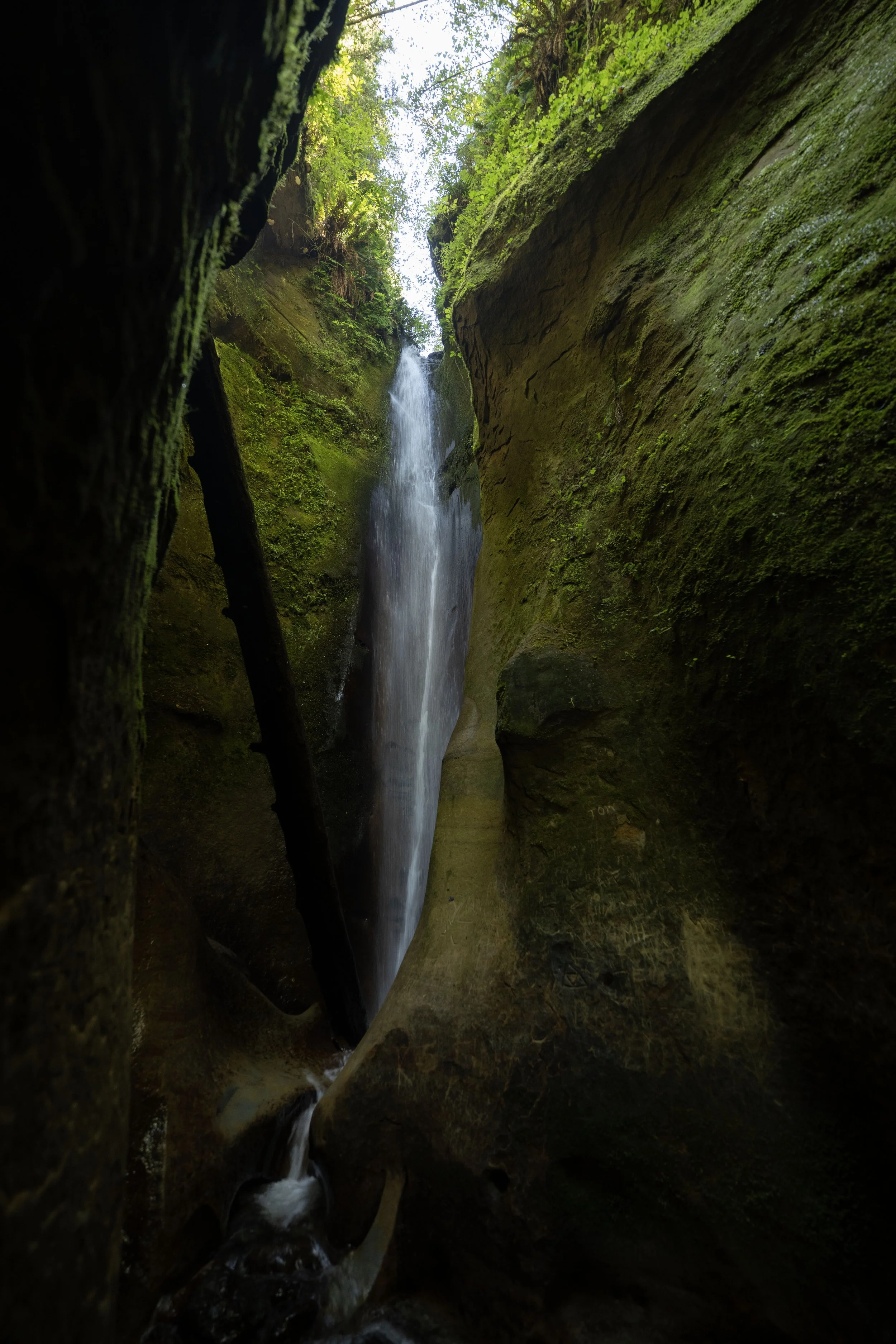 A narrow canyon with green moss-covered rocks and a waterfall cascading between the rocks, viewed from below.