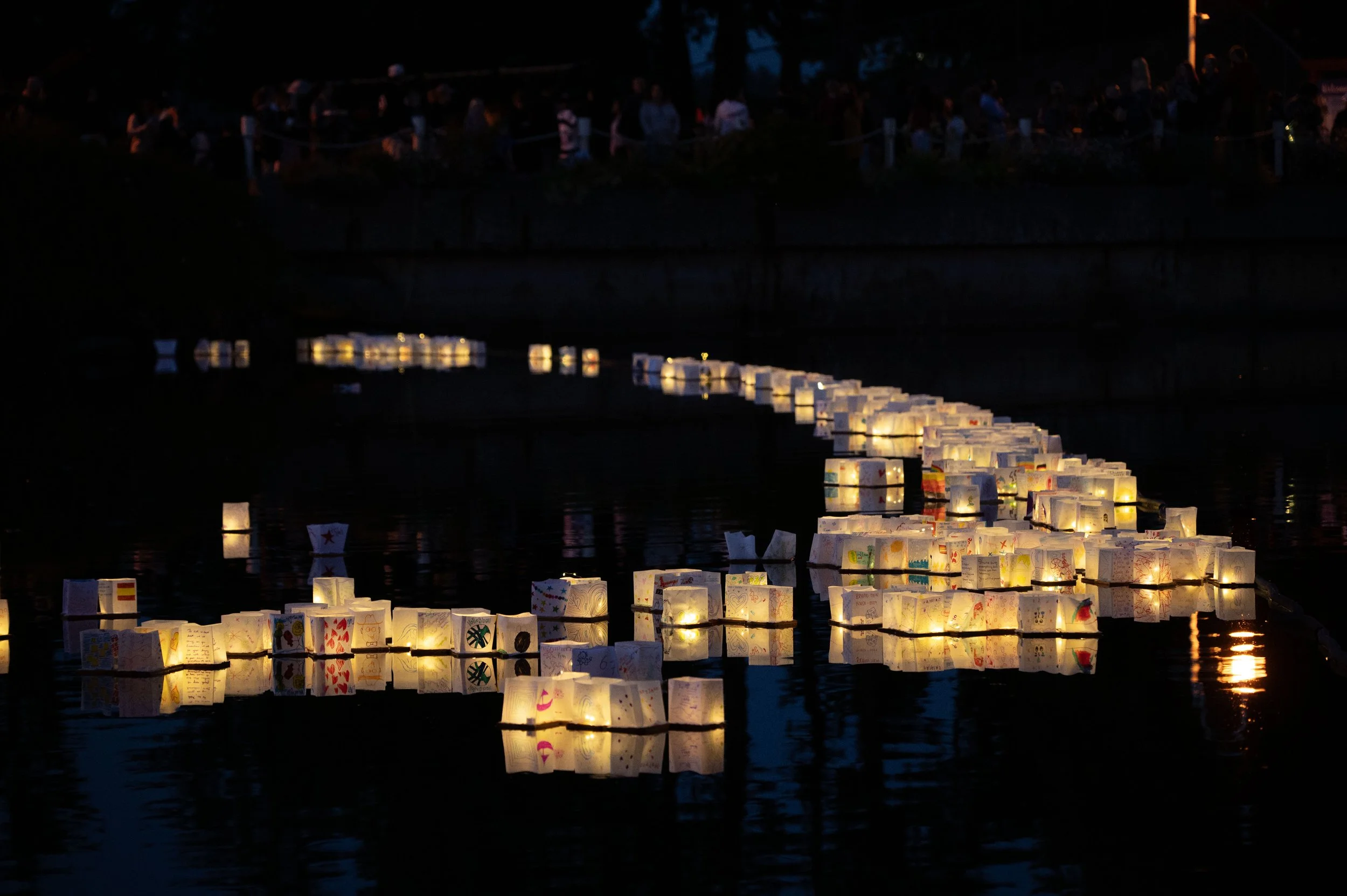 Numerous paper lanterns with drawings and messages float on a dark body of water during nighttime, creating a glowing display.