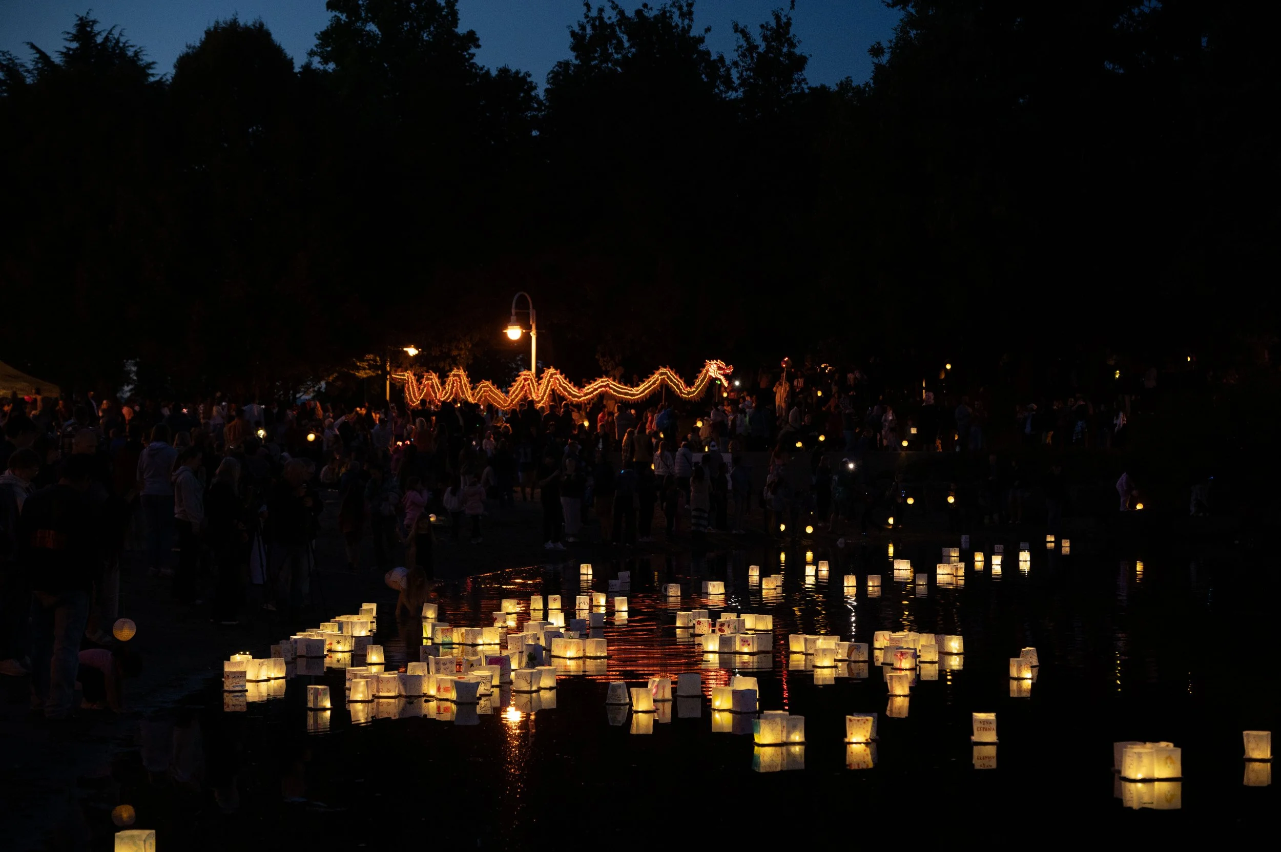 People gathered around a river during a festival at night, with floating lanterns on the water and illuminated dragon-shaped decorations in the background.
