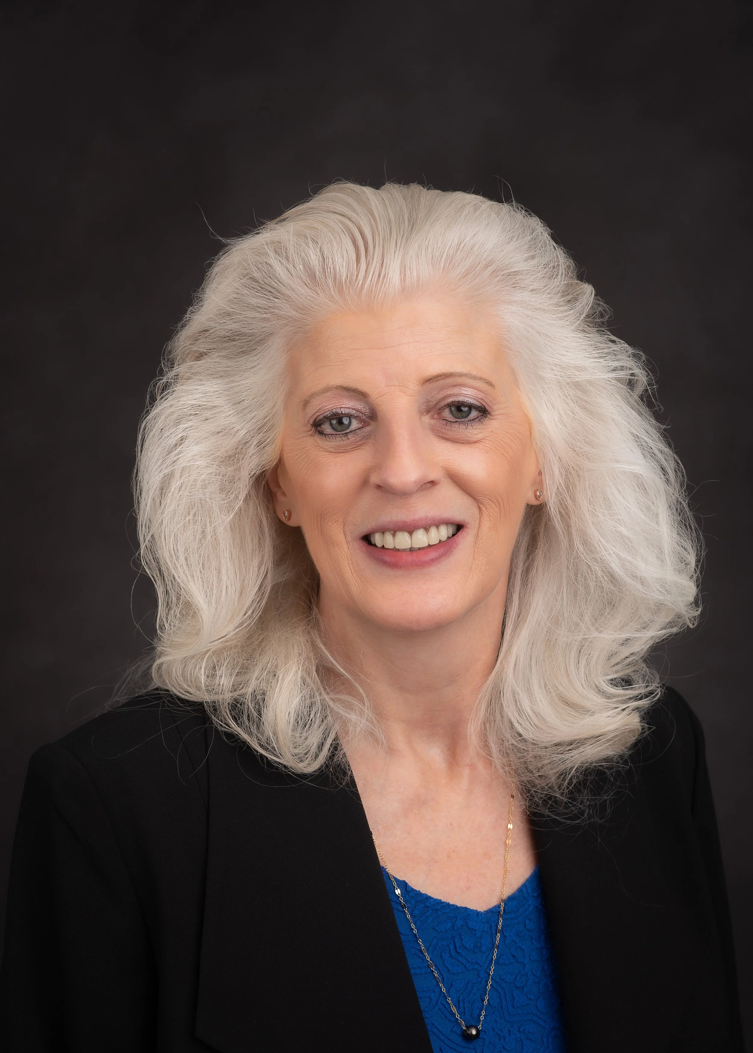Portrait of an elderly woman with long white hair, wearing a black blazer and a blue lace top, smiling against a dark background.