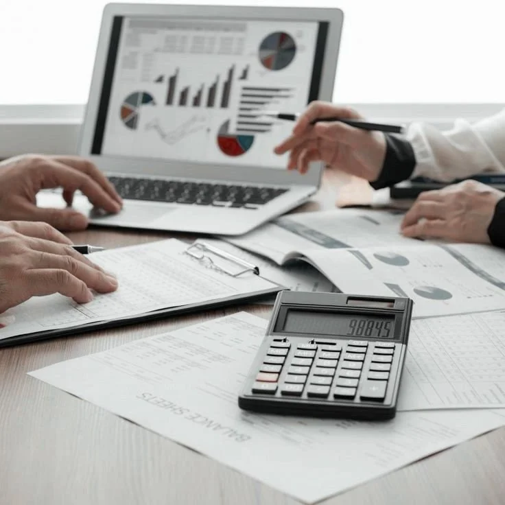 Business people working on financial reports with a laptop displaying graphs, a calculator, open notebooks, and documents on a desk.