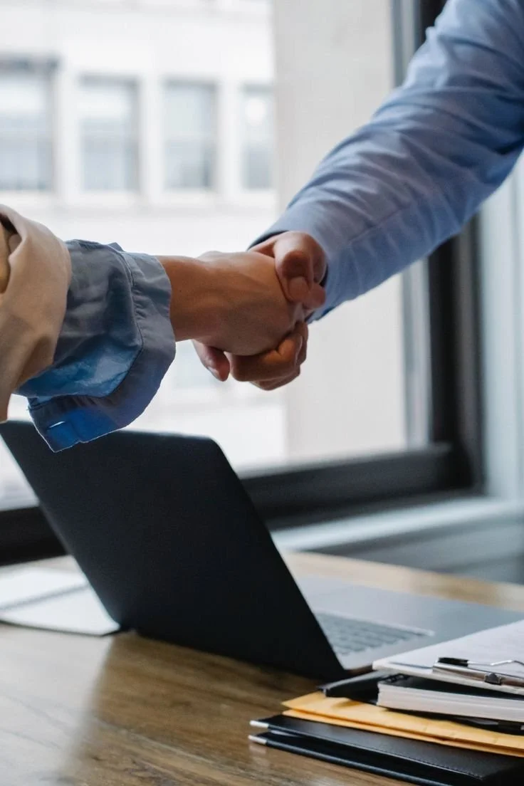 Two people shaking hands over a desk with a laptop and stacks of documents in an office setting.