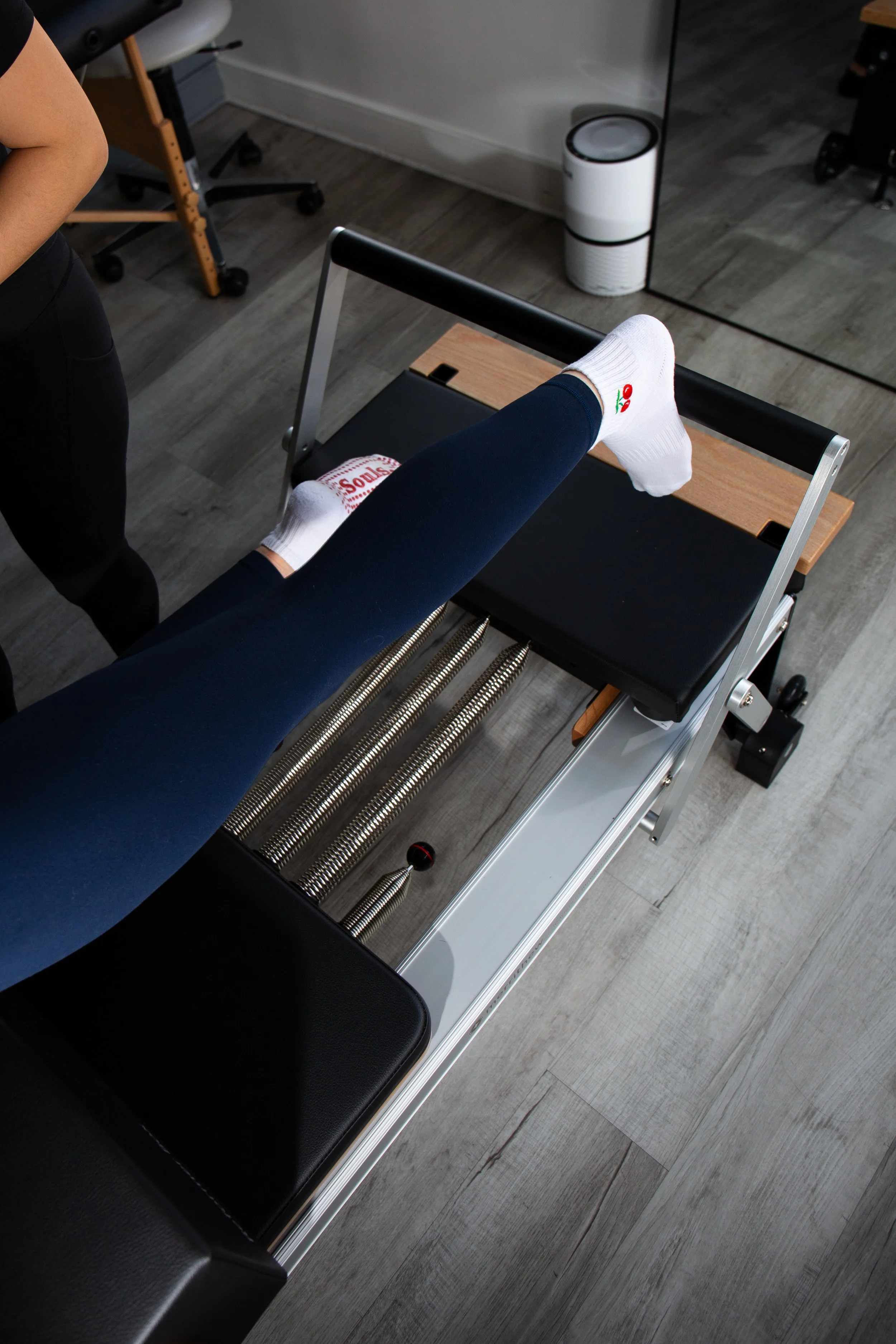 A person's leg with a white sock featuring a cherry design, resting on a black medical examination table in a clinical setting, with a wooden footrest and medical equipment in the background.