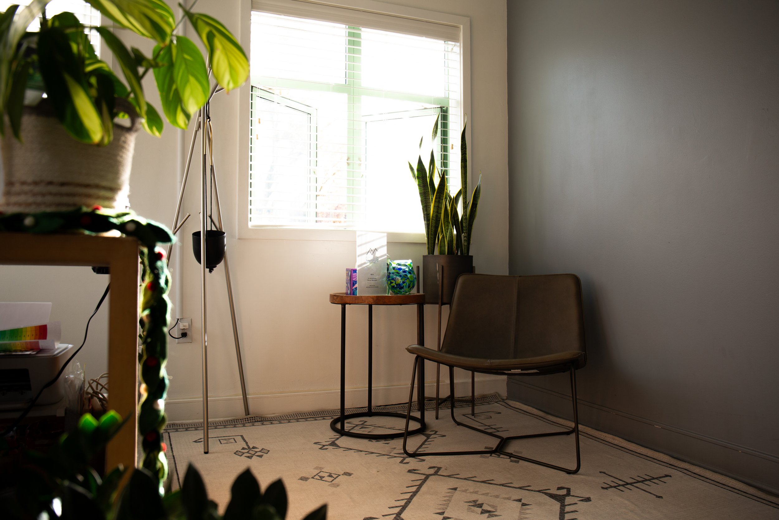 A cozy indoor corner with a window, potted plants, a small round wooden table with decorative items, and a brown chair on a patterned rug.