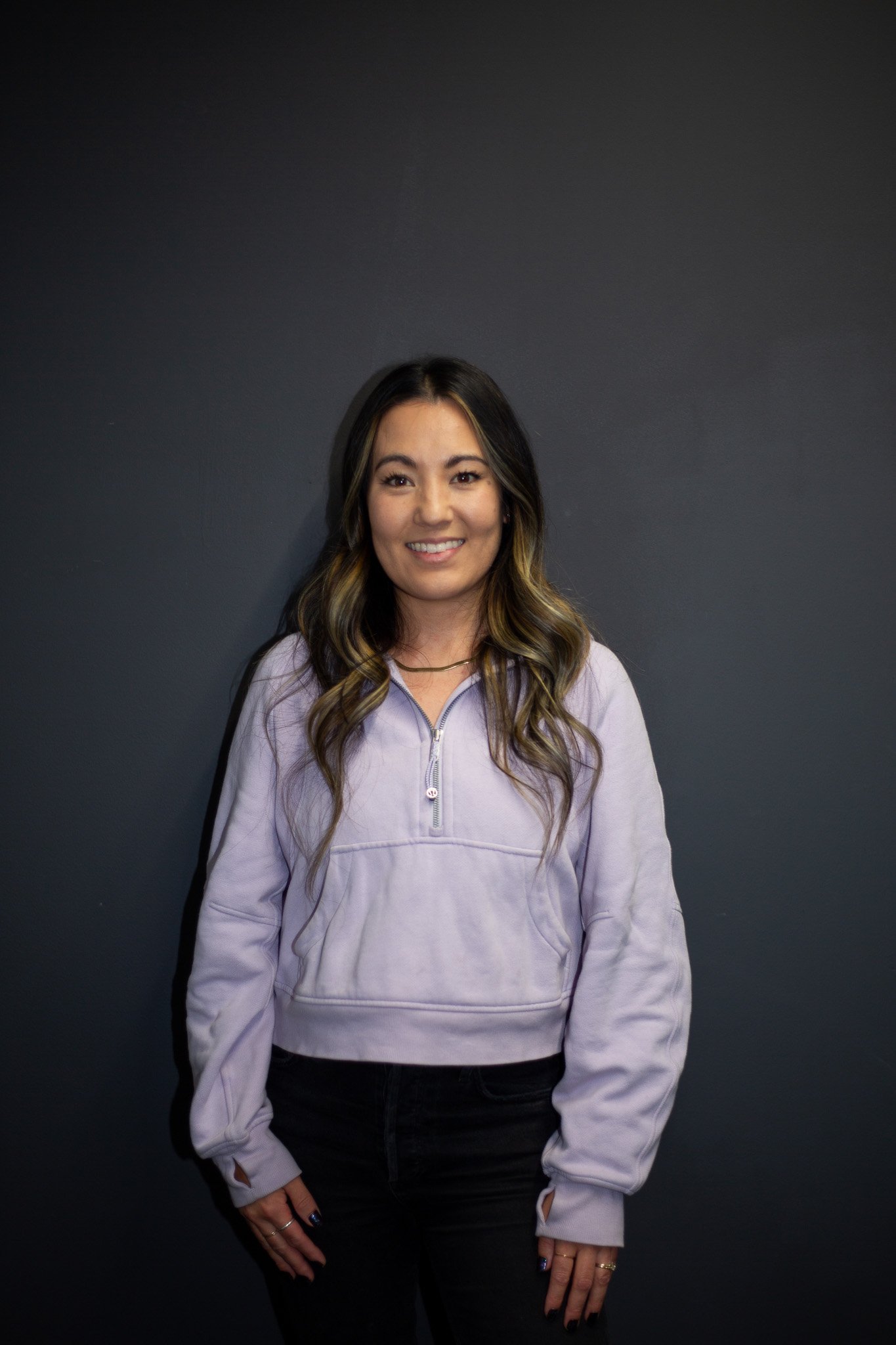 A woman with long wavy hair wearing a light purple hoodie and black pants, standing against a dark gray background and smiling at the camera.