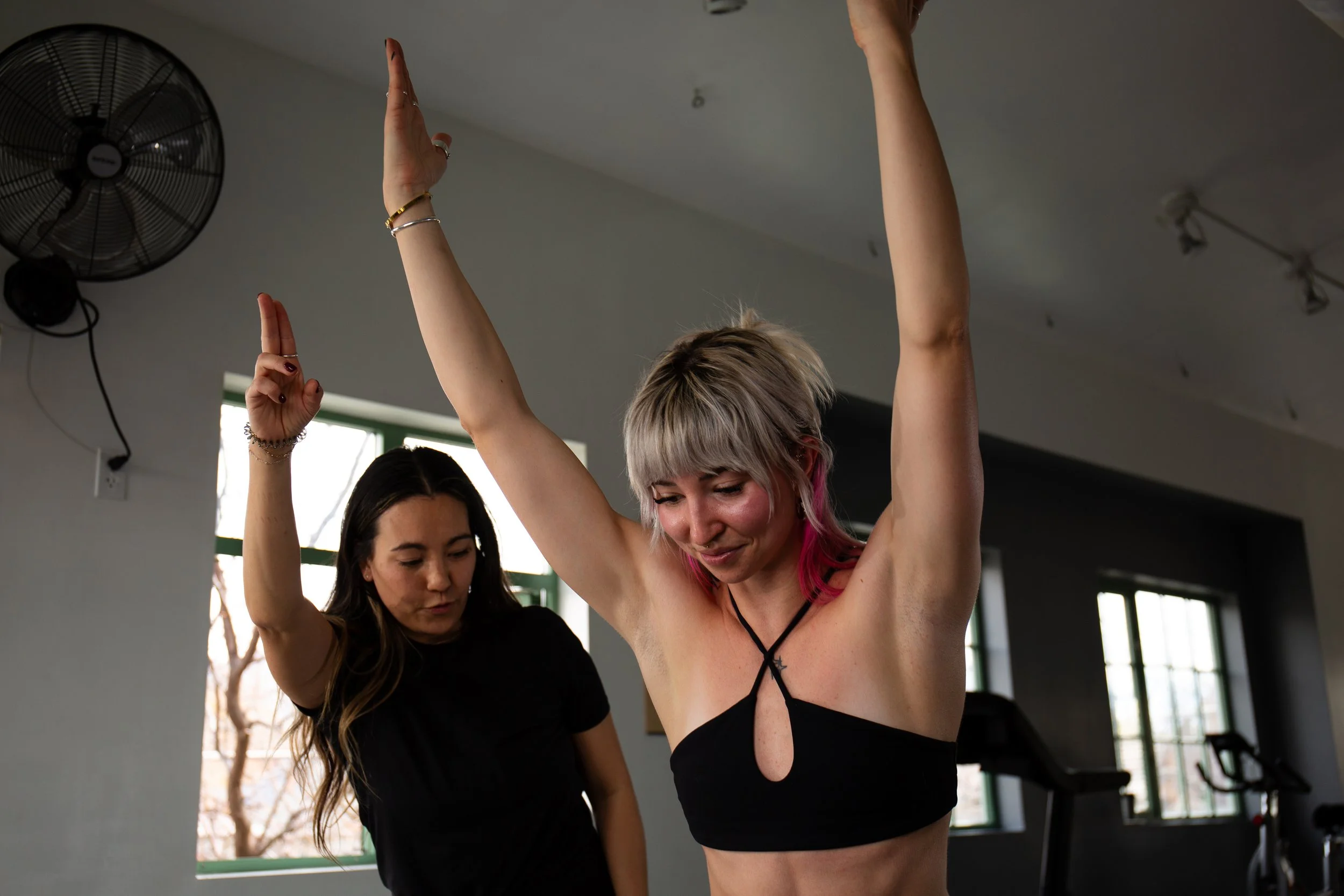 Two women are practicing yoga indoors; one with light hair and pink tips is smiling and raising her arms, while the other woman with dark hair is assisting her, standing nearby.