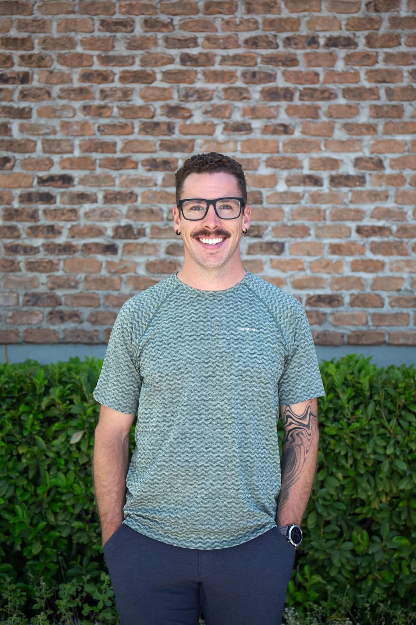 A smiling man with curly hair, glasses, a mustache, and earrings, standing outdoors in front of a brick wall with green shrubbery, wearing a gray patterned t-shirt and dark pants.