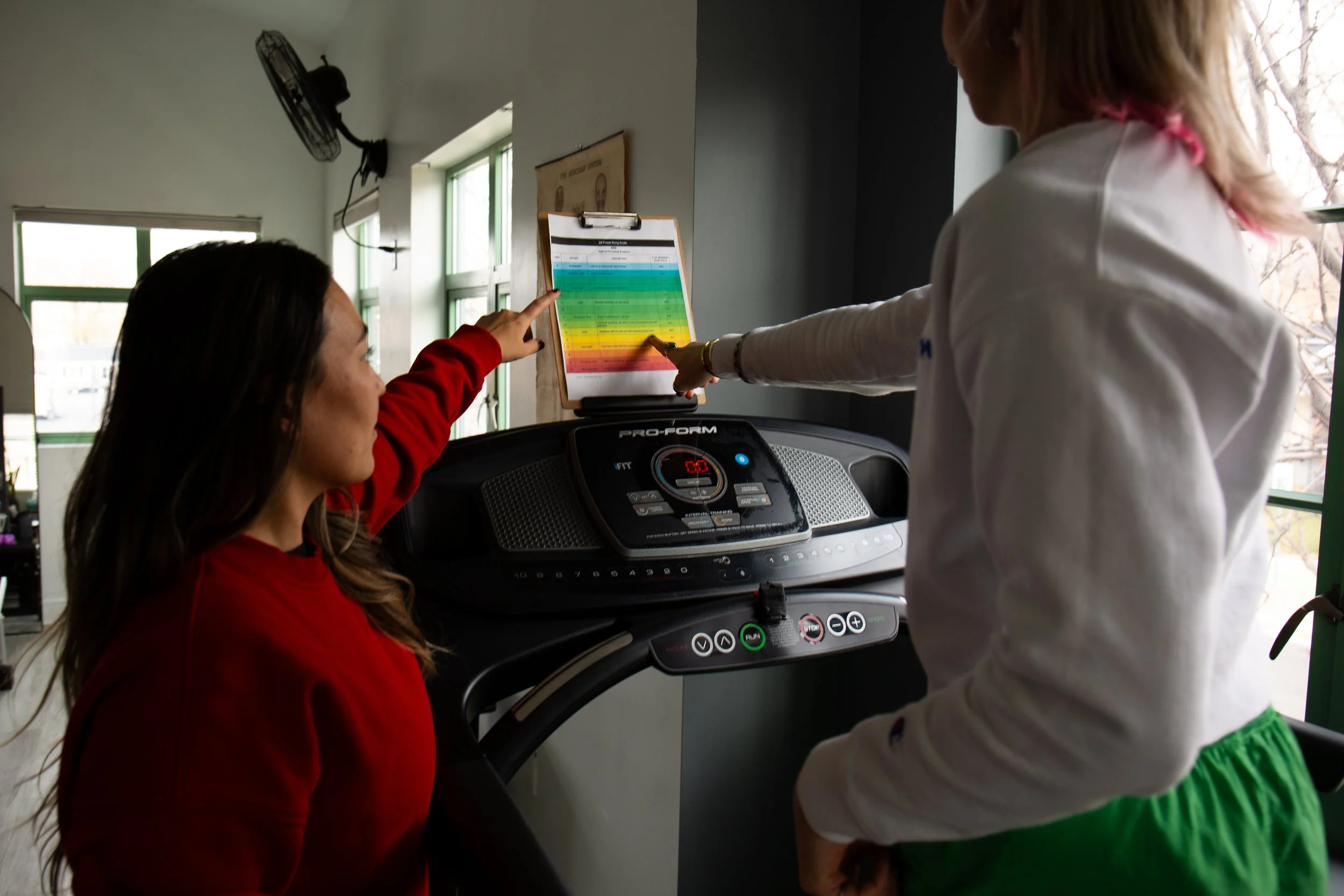 Two women at a treadmill, with one woman pointing at a colorful chart on a clipboard, in a room with windows and a wall-mounted fan.