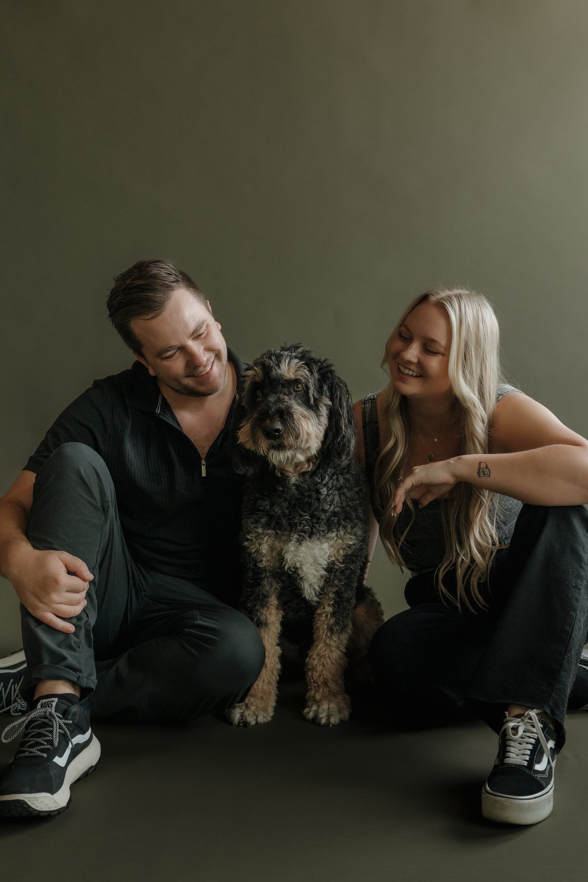 A smiling man and woman sitting on the floor with a large, black and gray dog between them. The man is wearing a black zip-up shirt, black pants, and sneakers, and the woman has long blonde hair, a sleeveless top, and tattoos. The dog appears to be a mixed breed, with a curly coat and expressive brown eyes. They are all looking at each other and seem happy.