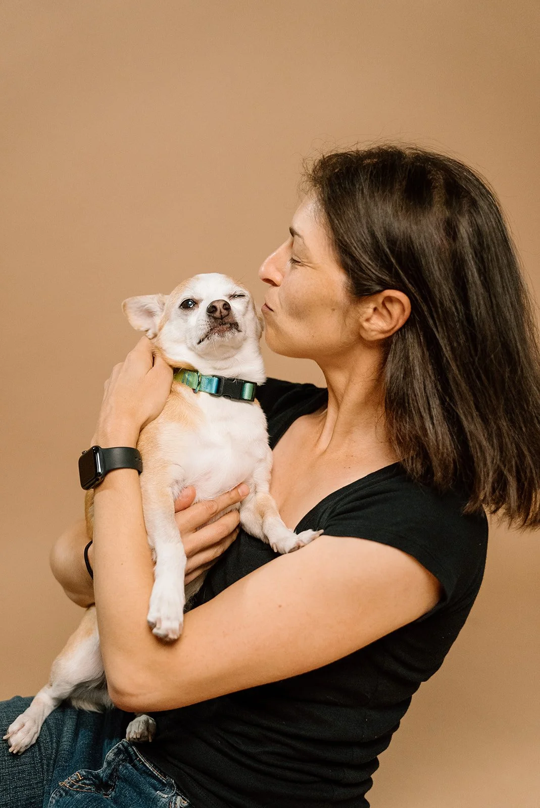 A woman holding a small dog close, about to kiss it, against a plain beige background.
