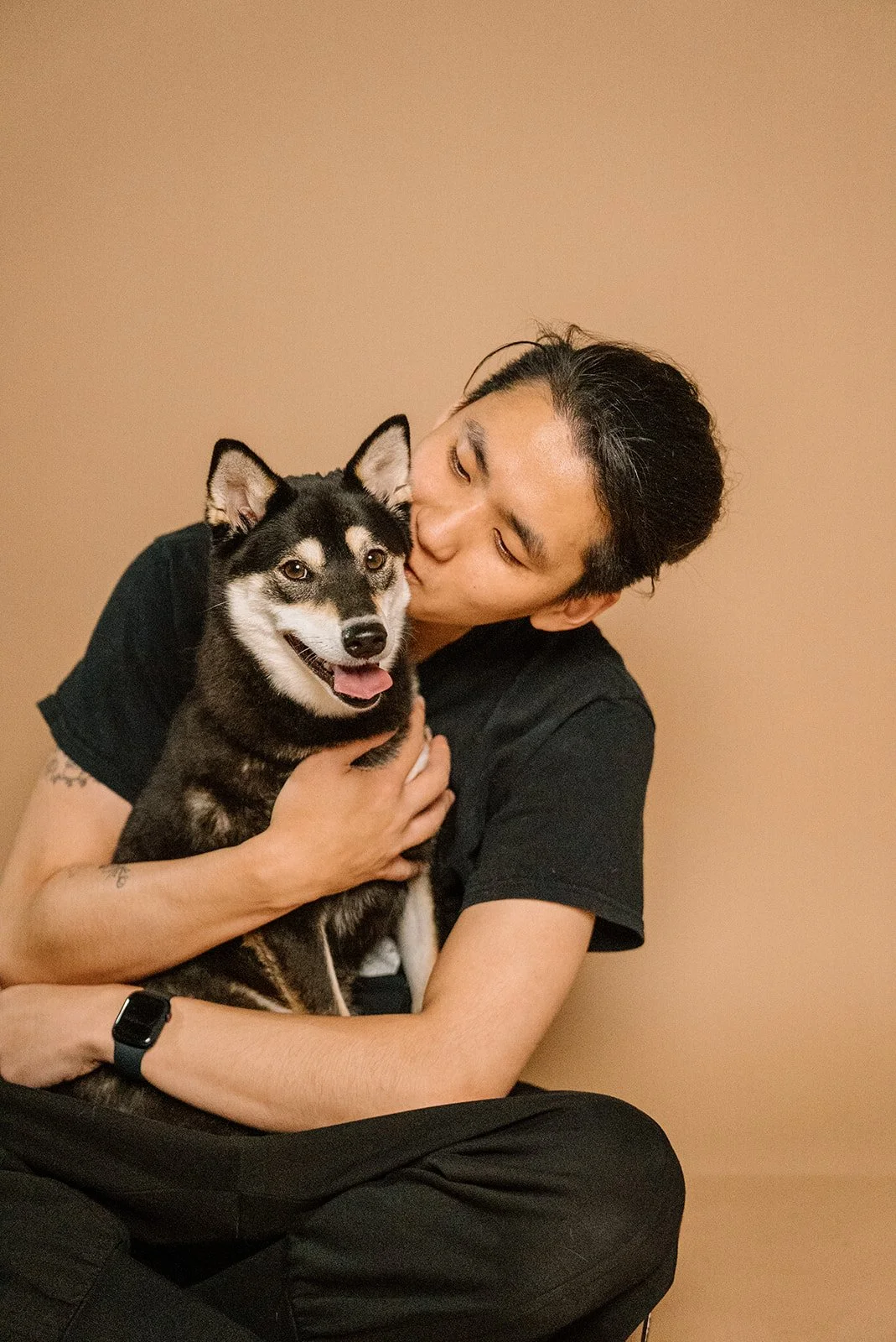 A person with black hair wearing a black shirt, sitting cross-legged, hugging and kissing a small husky dog with black and white fur, against a beige background.