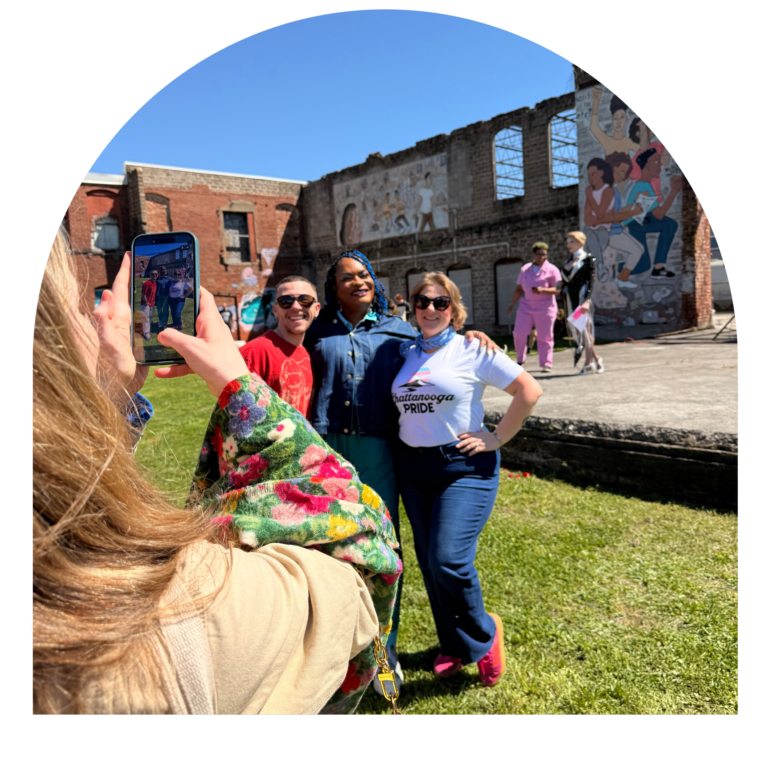 A group of diverse people posing for a photo outdoors during the Chattanooga Pride event, with some individuals smiling and one person holding a smartphone to capture the moment. The background includes murals on an old brick building and a clear blue sky.