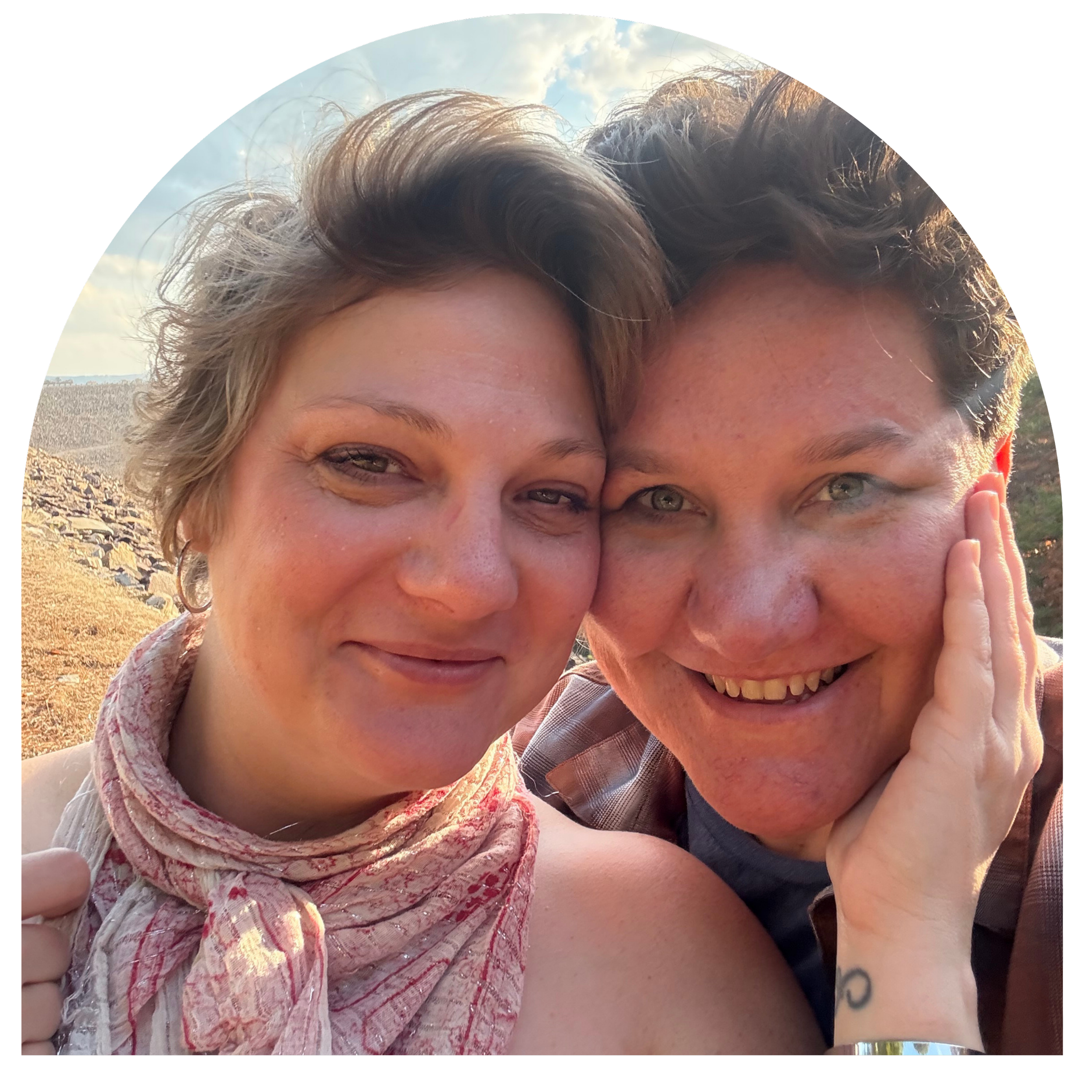 Close-up of two women smiling and enjoying an outdoor setting with clear sky, rocky terrain, and a distant landscape in the background.