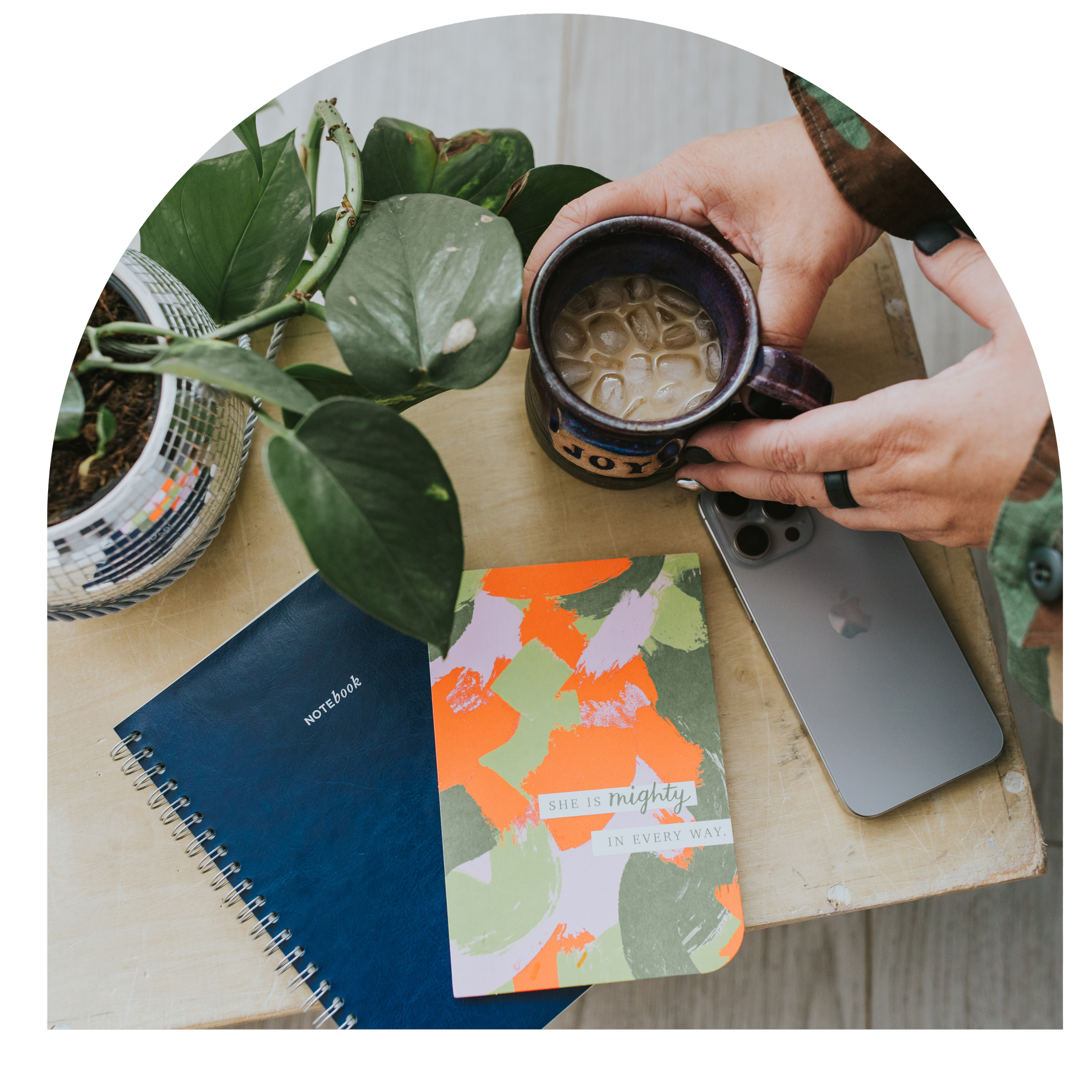 Interior scene with potted plants, a ceramic mug with iced coffee, notebooks, and a smartphone on a wooden surface.