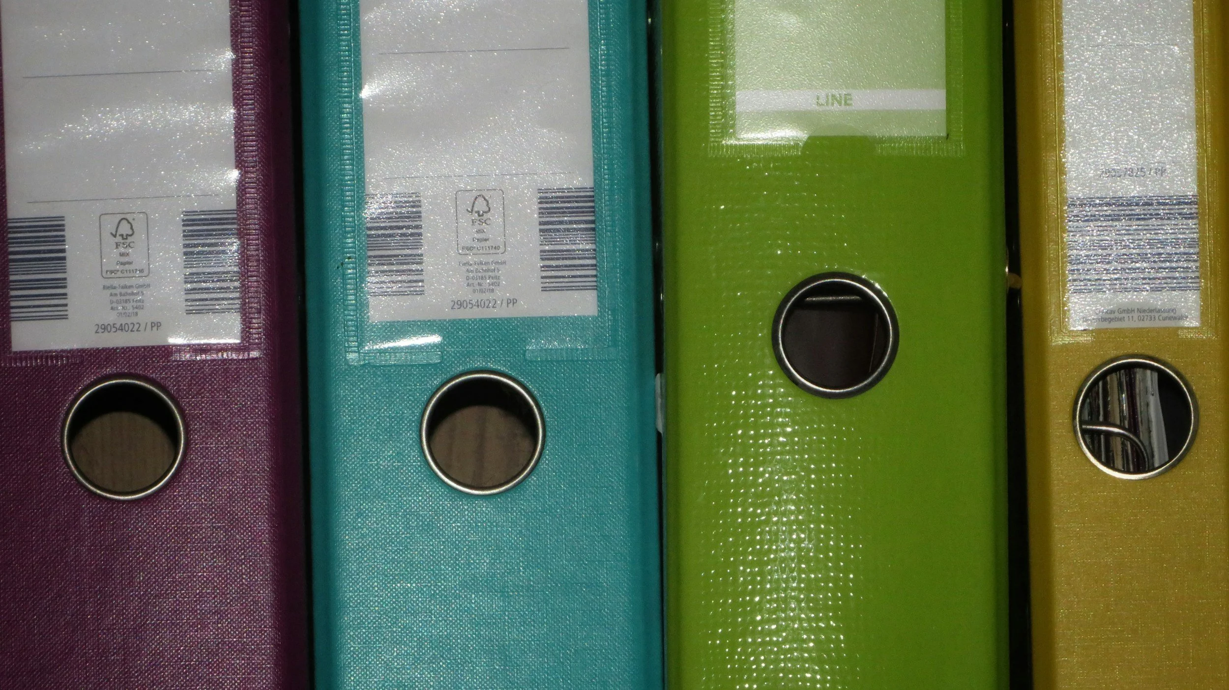 Colorful binders in purple, blue, green, and yellow placed side by side, with circular label holes and barcodes on the spines.