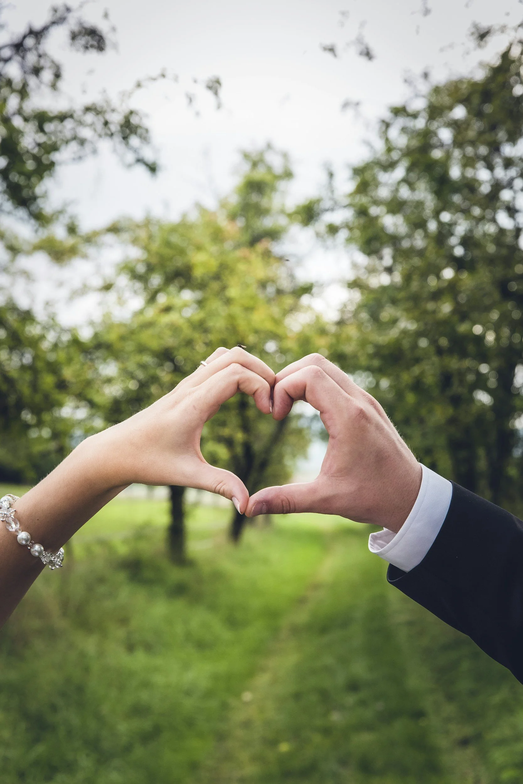 Two hands, one from a woman wearing a bracelet and one from a man in a suit, form a heart shape outdoors with green trees and grass in the background.
