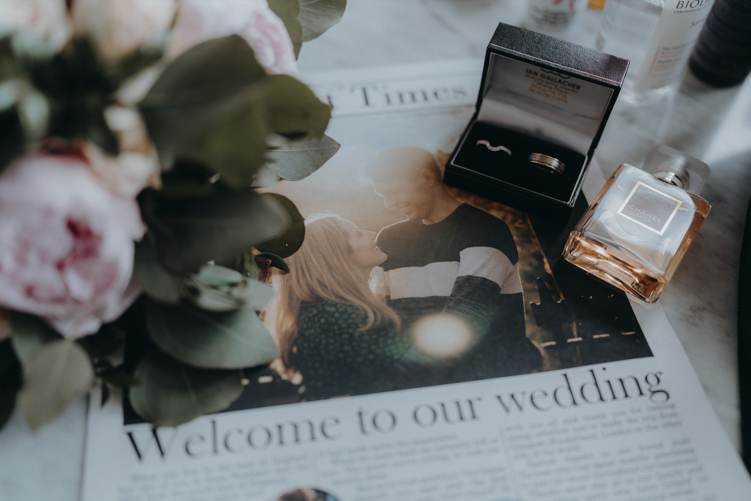 The front page of a wedding newspaper with a photo of a bride and groom. Surrounding it are flowers, a pair of wedding rings in a black box, and a Chanel perfume bottle.