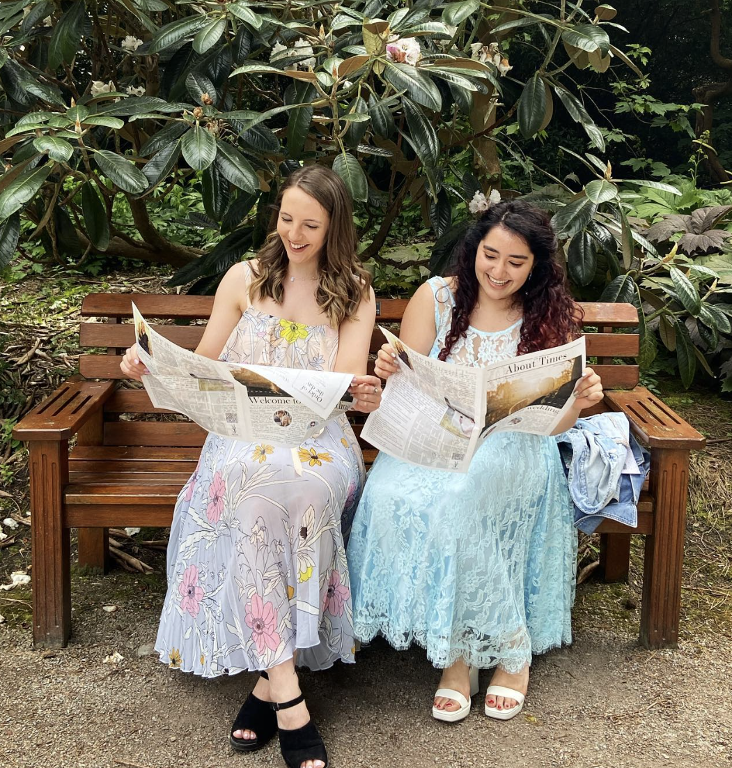 Two wedding guests sitting on a bench and reading personalised wedding newspapers.