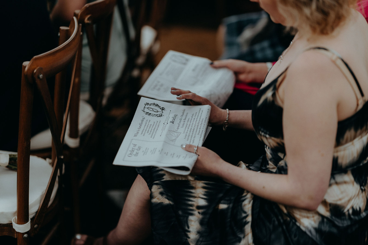 A female wedding guest, sitting on a wooden chair, reading a wedding newspaper.