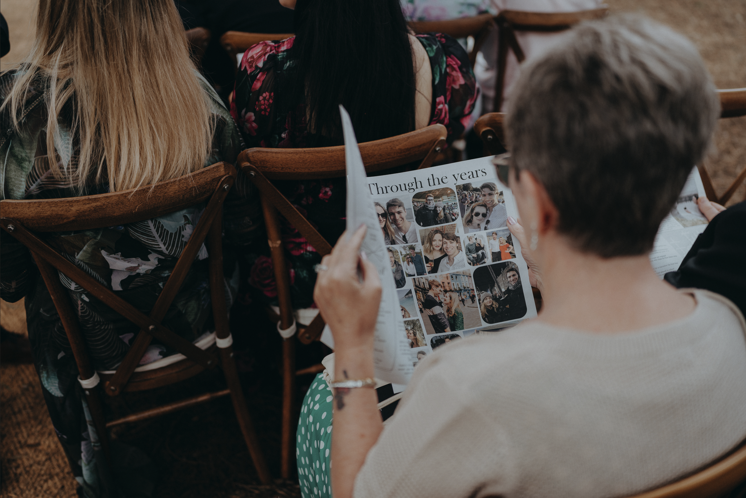 A woman with short gray hair reading a wedding newspaper containing a photo collage called "Through the years" at an outdoor wedding ceremony.