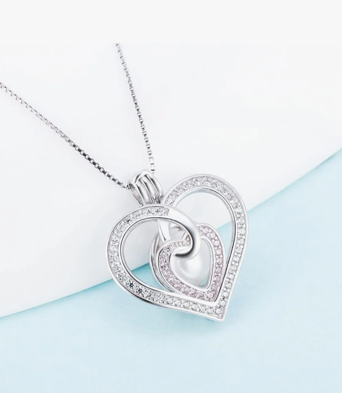 Silver necklace with two intertwined heart-shaped pendants, decorated with small sparkling stones, resting on a light blue and white surface.