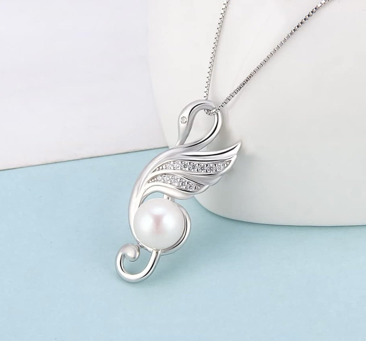 Silver necklace with a swan-shaped pendant, featuring a large pearl and small embedded rhinestones, hanging against a white and light blue background.