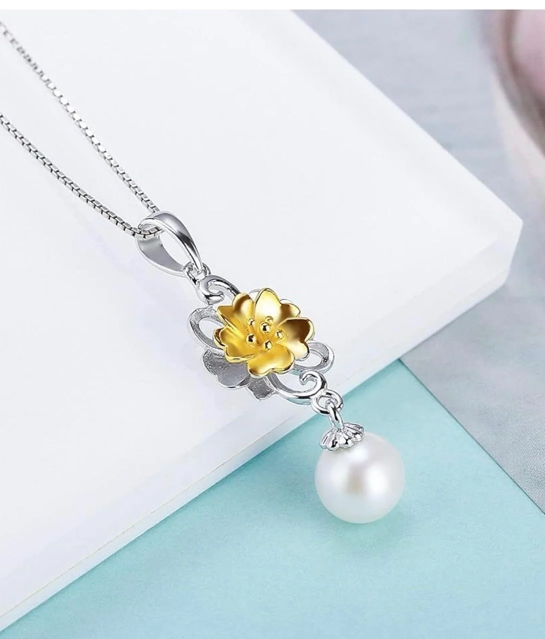 A silver necklace with a yellow flower pendant, a silver swirl detail, and a large white pearl hanging at the bottom, placed on a white and light blue surface.