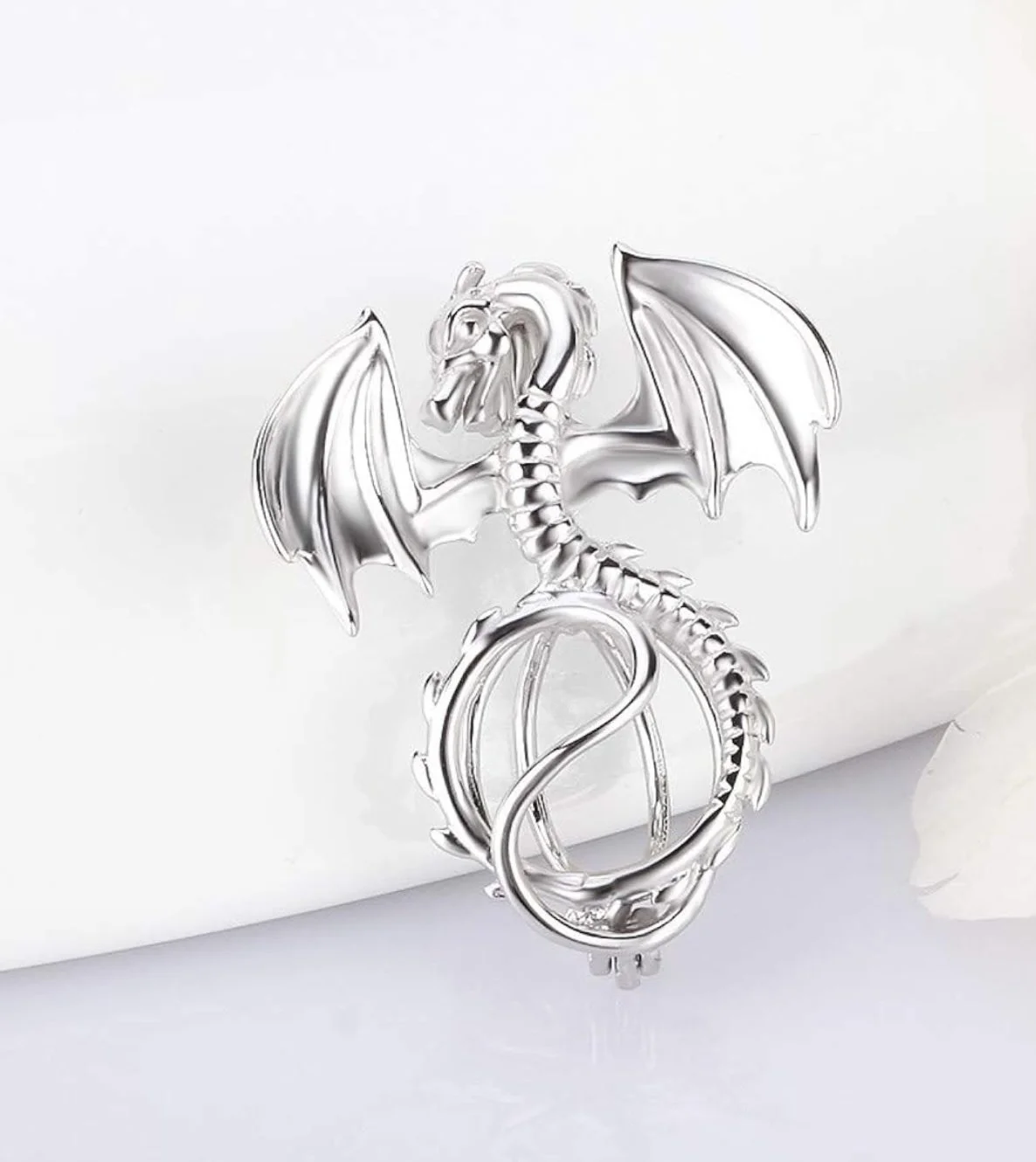 Silver metallic pendant in the shape of a dragon with wings, a long tail, and a spherical element at the bottom.